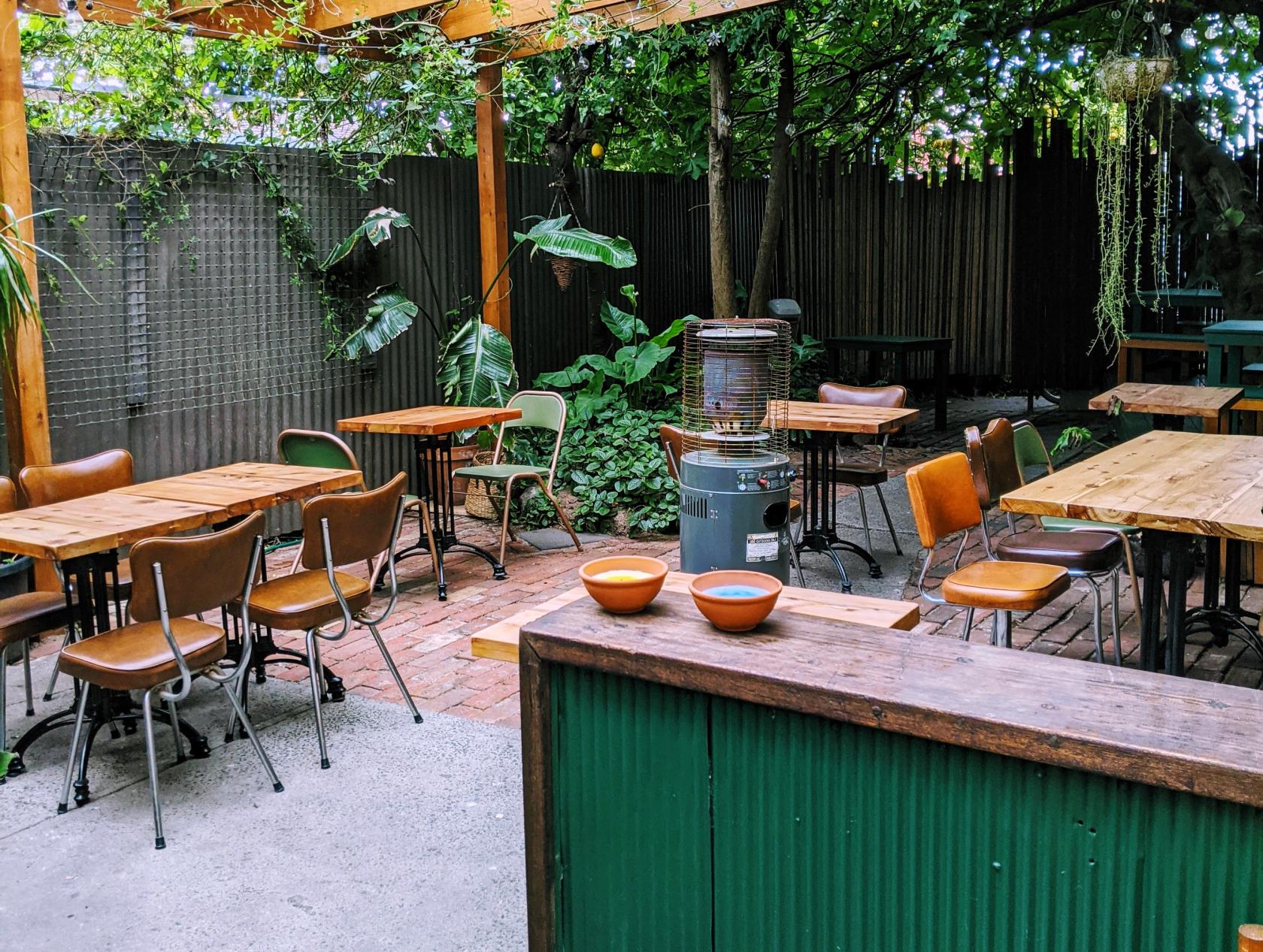An outdoor bar with wooden tables, mixed chairs, leafy plants, and a patio heater at Norte Wines.