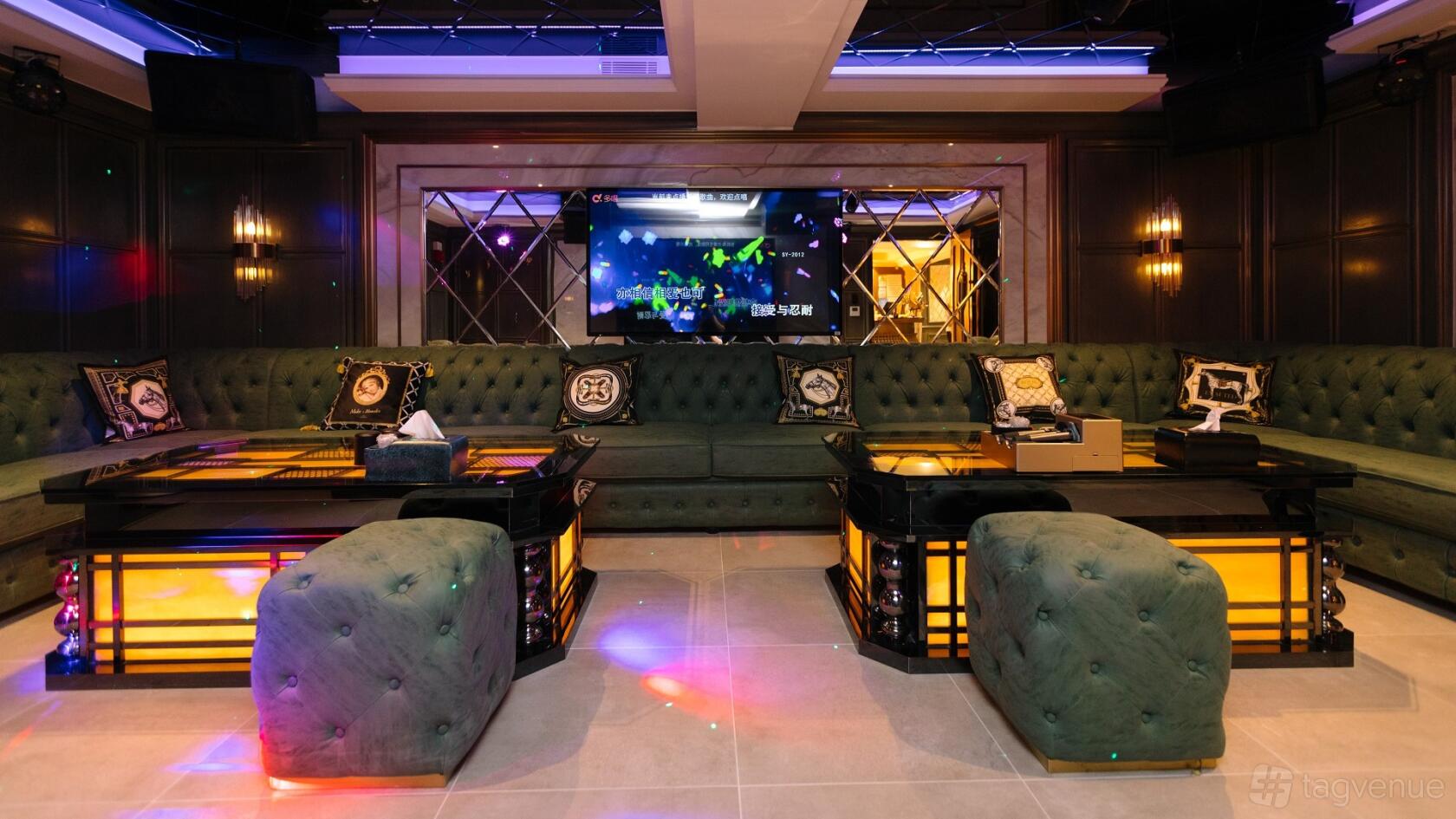 A karaoke event space with a long green sofa, illuminated tables, plush ottomans, and a large screen at Sugar karaoke.