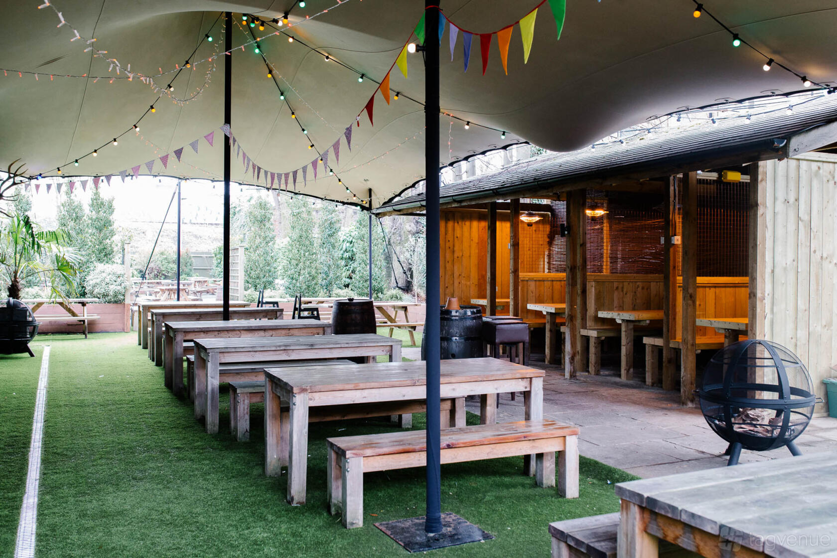An outdoor pub area with wooden benches, a canopy, string lights, and artificial grass at The Glasshouse.