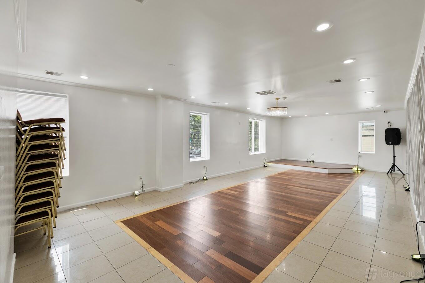 A hall with a small wooden stage, tile and wood flooring, stacked chairs, and large windows at Haven Hall Philly.