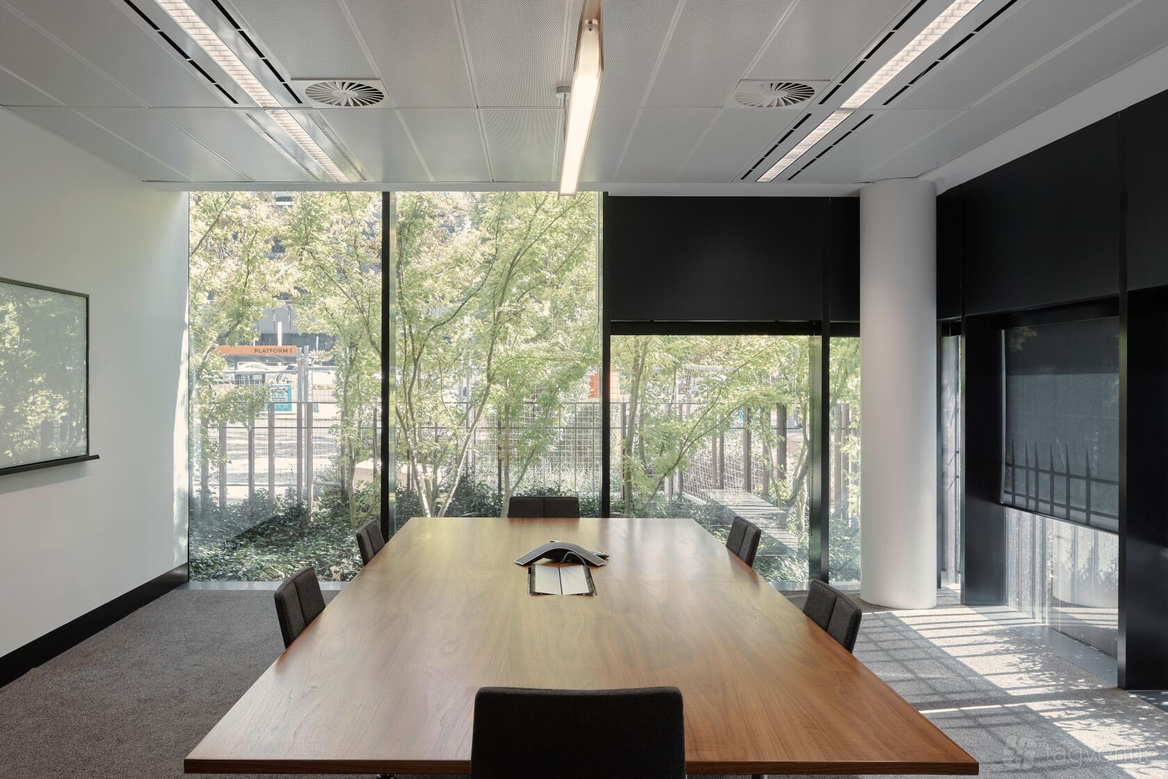 A meeting room with a large wooden table, black chairs, and floor-to-ceiling windows overlooking trees at Flex by ISPT at 4 National Circuit.