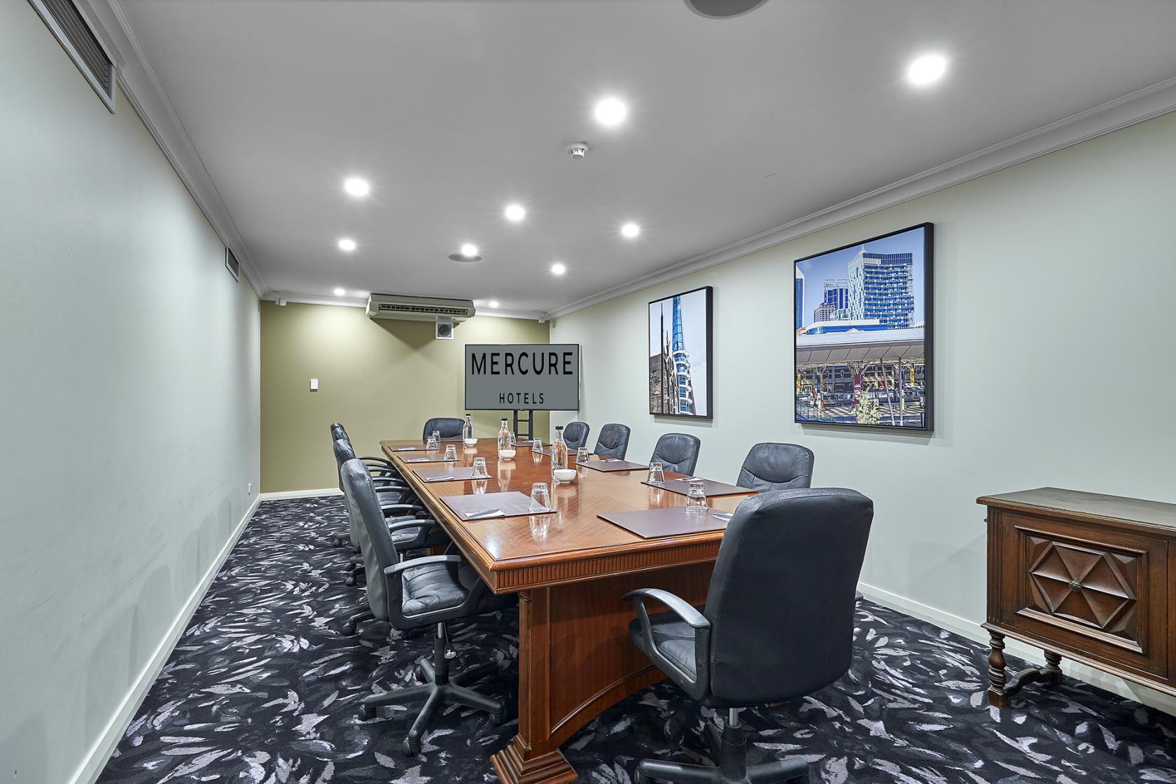 A boardroom with a long wooden table, black leather chairs, patterned carpet, and framed city prints at Mercure Perth on Hay.