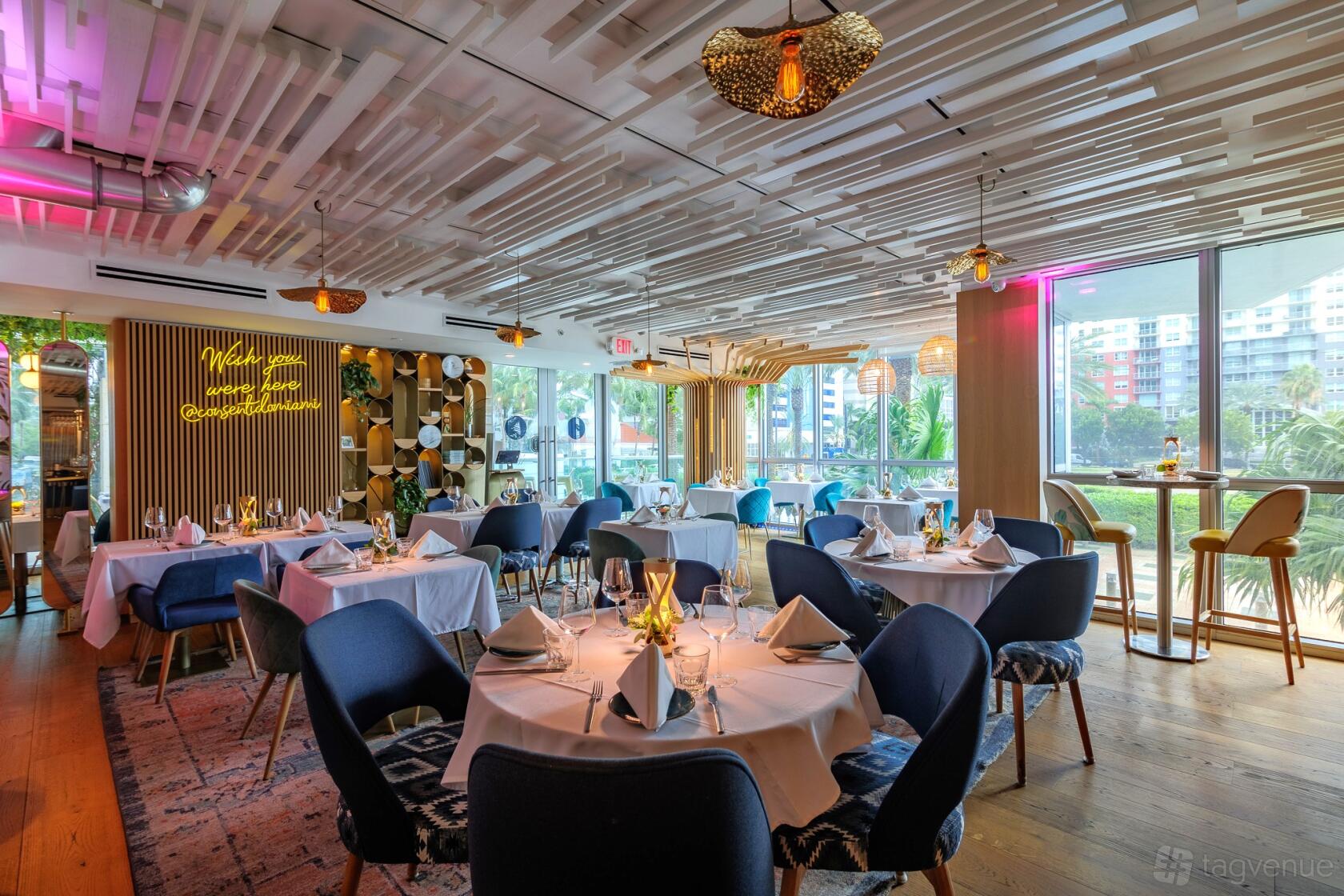 A restaurant dining room with large windows, round tables with white linens, and blue chairs at ConSentido Restaurant.