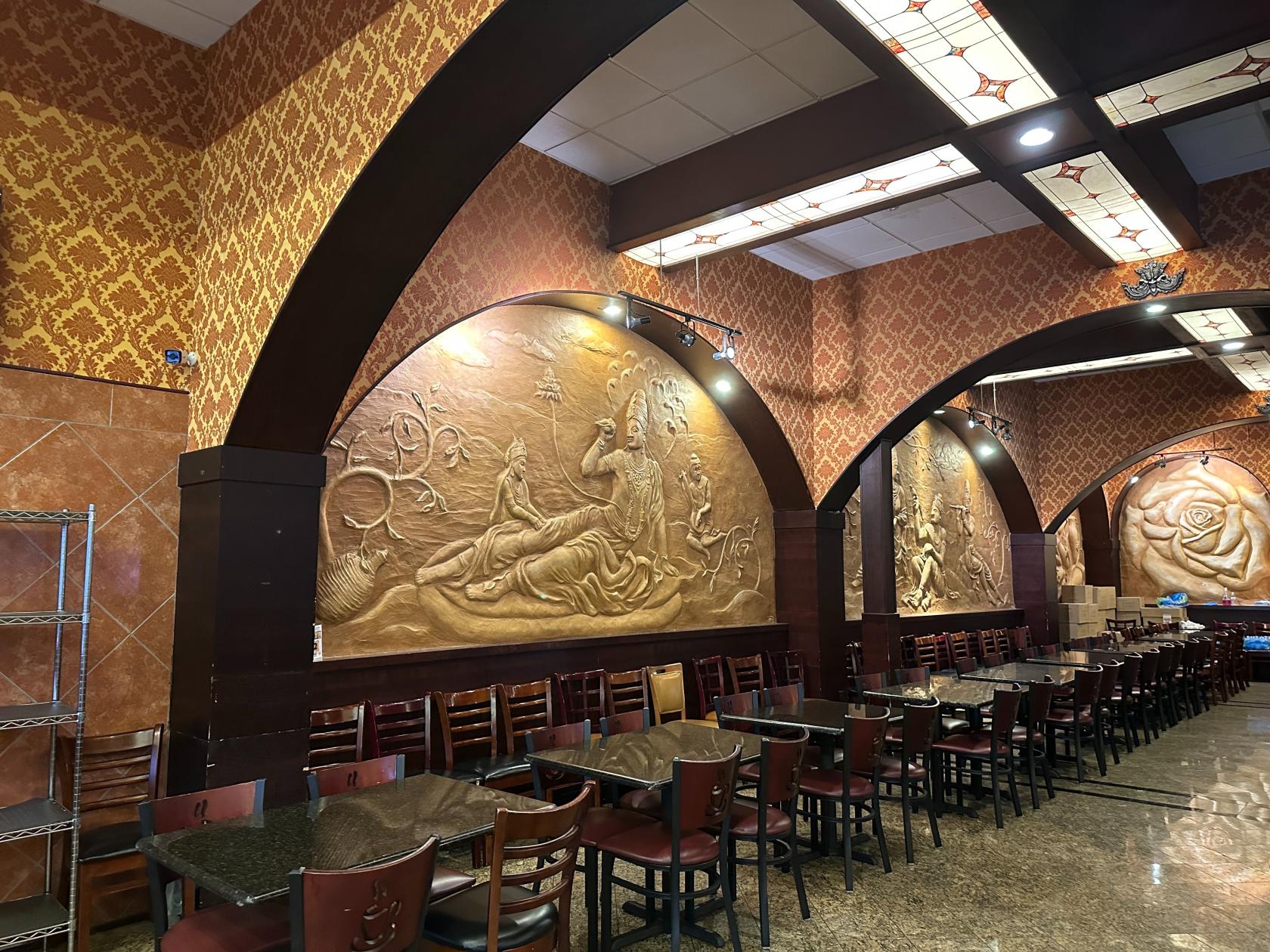 An event space in a restaurant with large carved wall murals, patterned wallpaper, and rows of dark wood tables at Chennai Café.