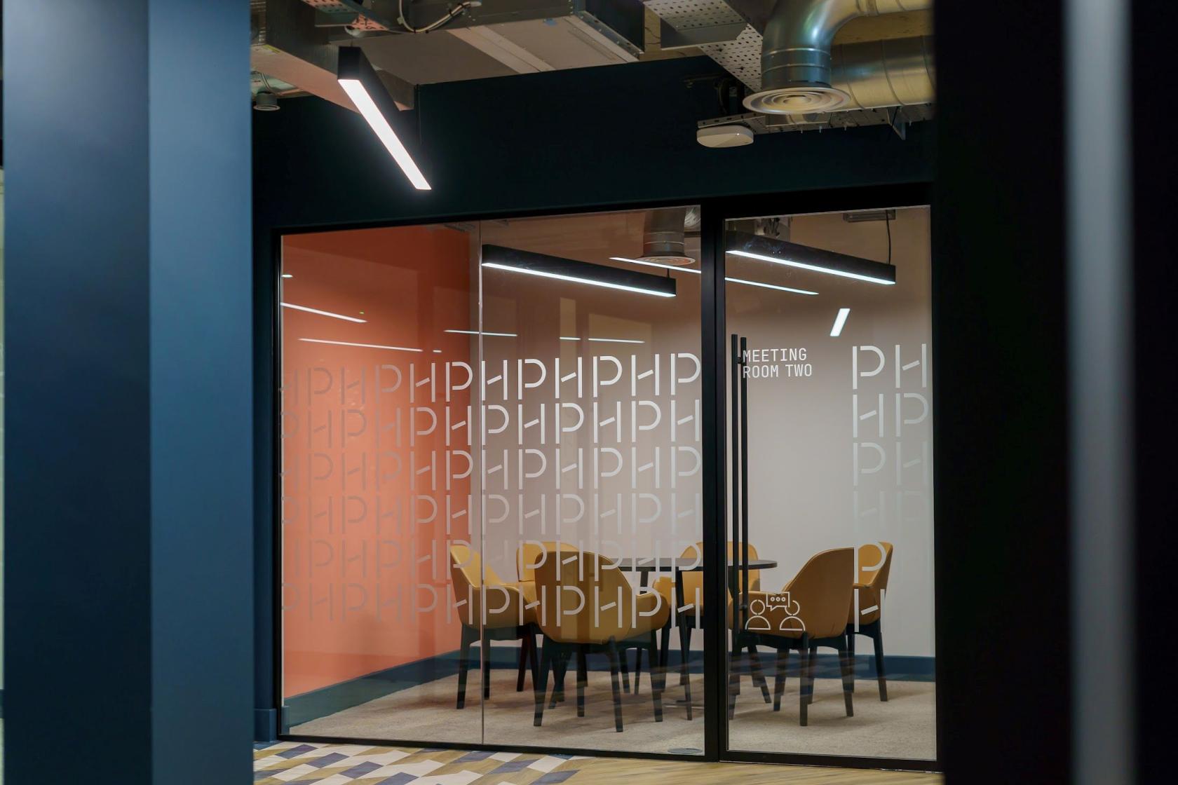 A meeting room with glass walls, round table, mustard chairs, and exposed ceiling at Wizu Workspace - Newcastle - Portland House.