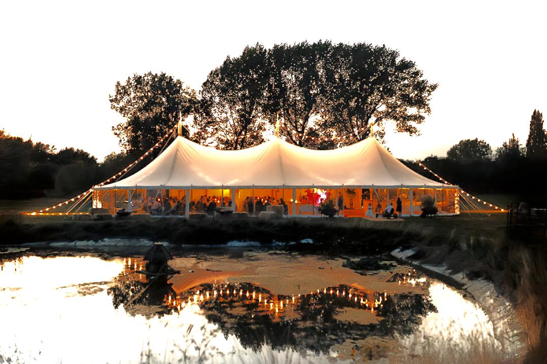 An outdoor event venue with a large marquee tent, string lights, and pond reflection at Peacock and Bow.