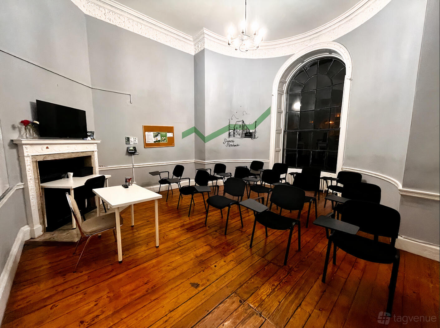 A classroom with black chairs, wooden floors, a large arched window, and a white fireplace at North Great George's Street.