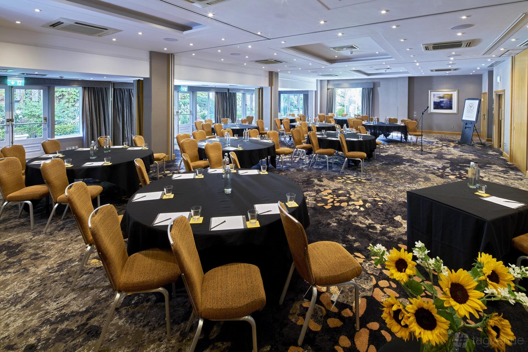 A hotel function room with round tables, tan upholstered chairs, floor-to-ceiling windows, and patterned carpet at The Bull Hotel.