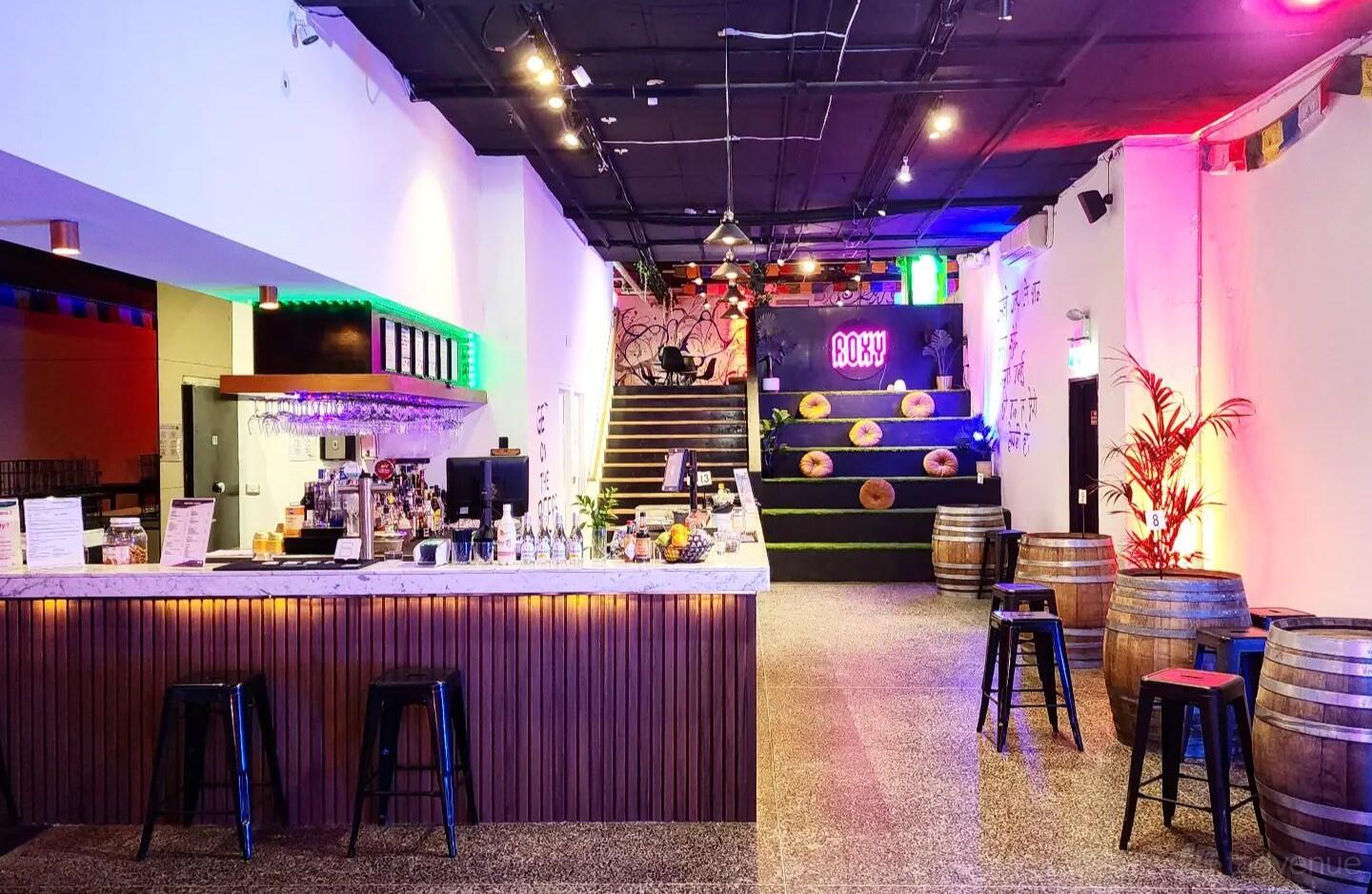 A restaurant with a marble bar counter, high stools, colorful accent lighting, and wooden barrels at Roxy Kitchen and Bar.