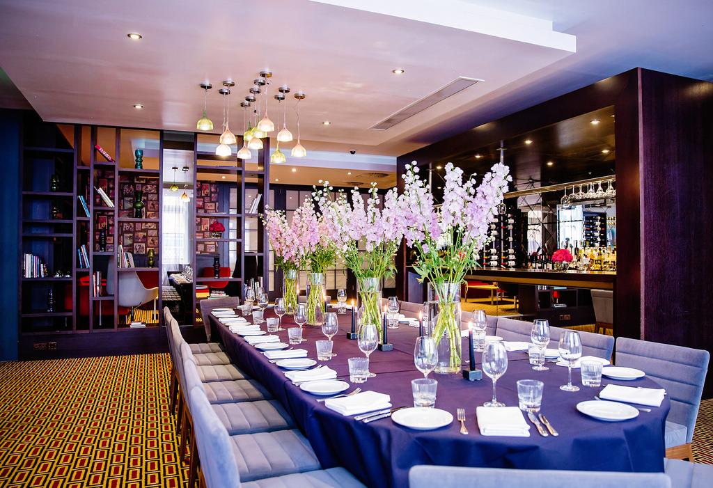 A hotel lounge with a long banquet table, tall floral centerpieces, glassware, and a mirrored bar at Malmaison London.