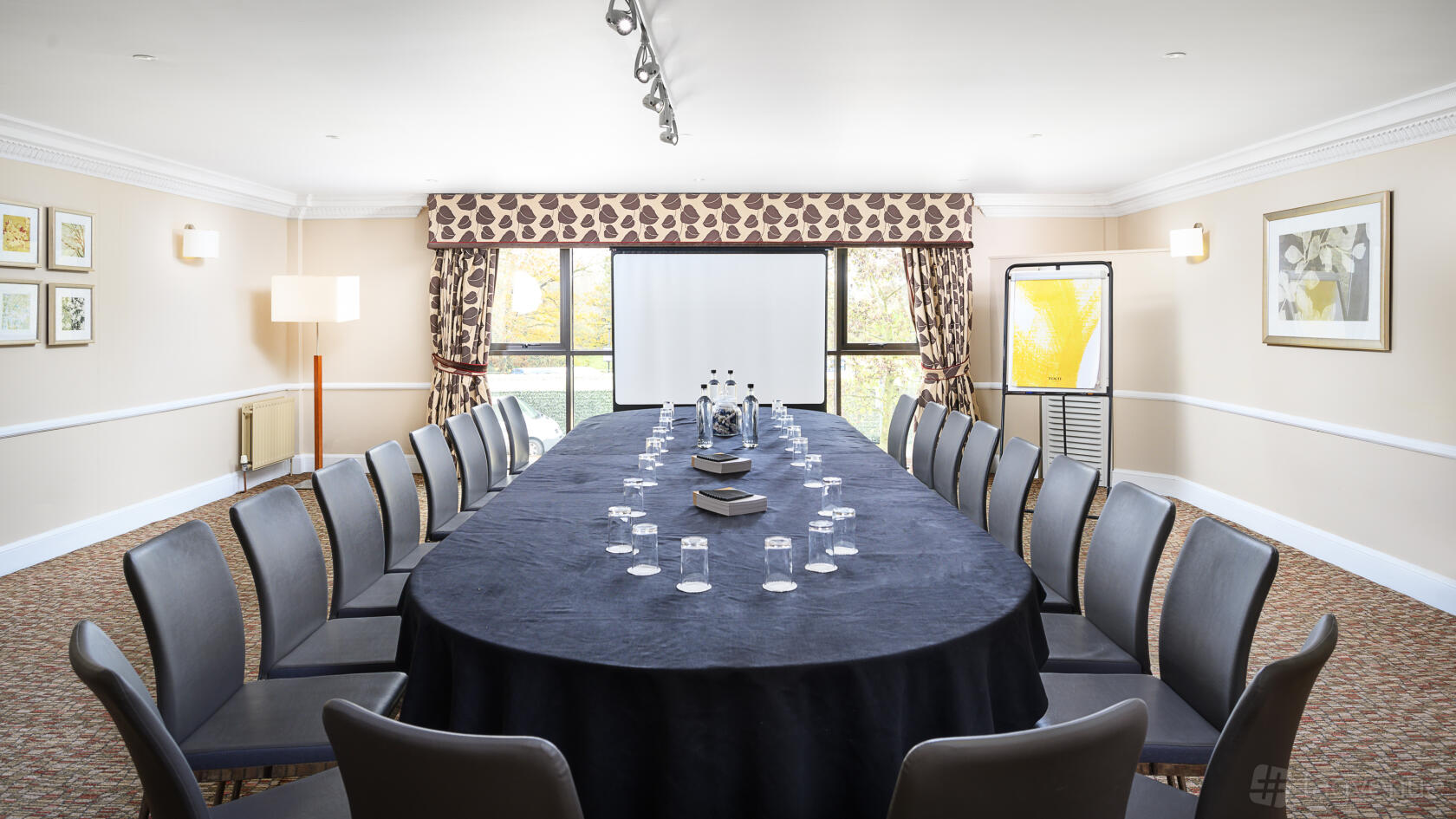 A meeting room with an oval conference table, grey chairs, and a projector screen at Voco Oxford Spires.