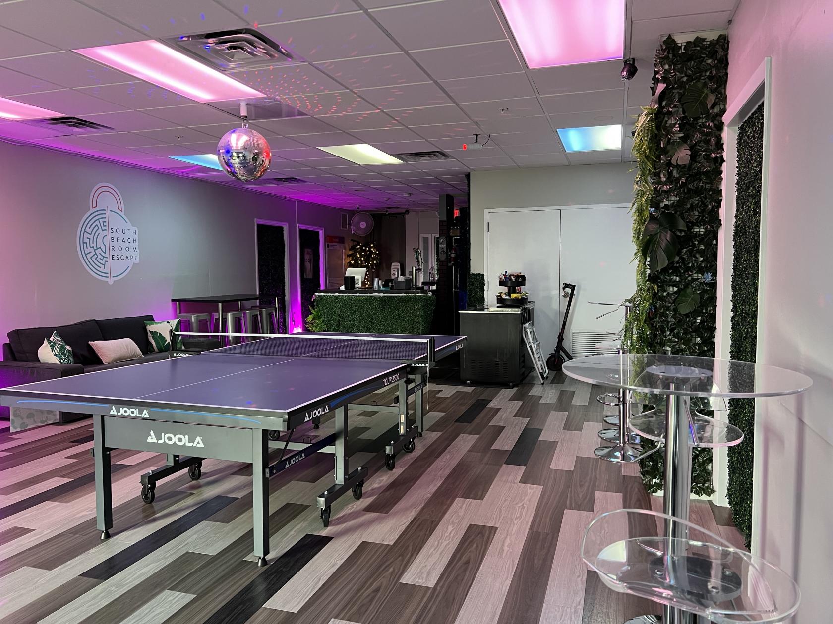 An indoor event space with a ping pong table, disco ball, lounge seating, and wall greenery at South Beach Room Escape Bar & Rooftop.