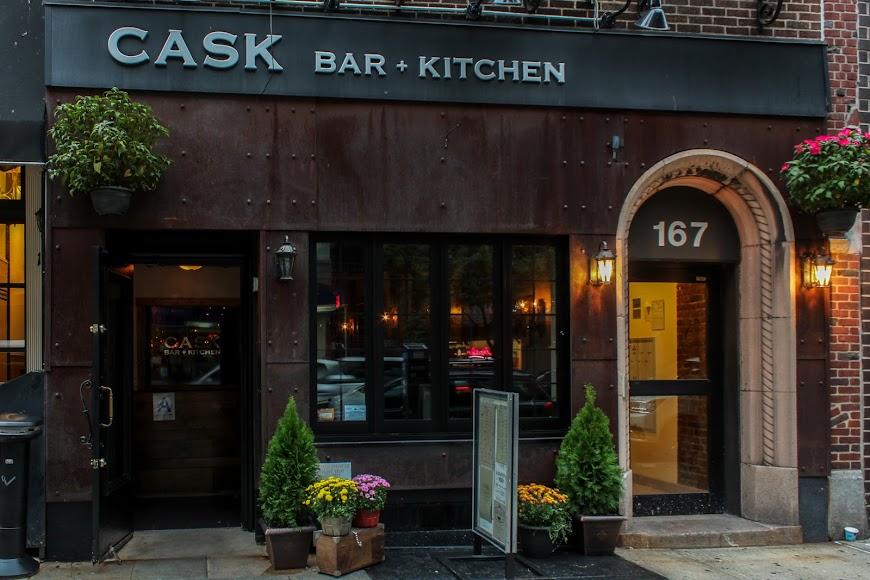 A pub with a dark wood facade, arched doorway, and potted plants at the entrance of Cask Bar + Kitchen.