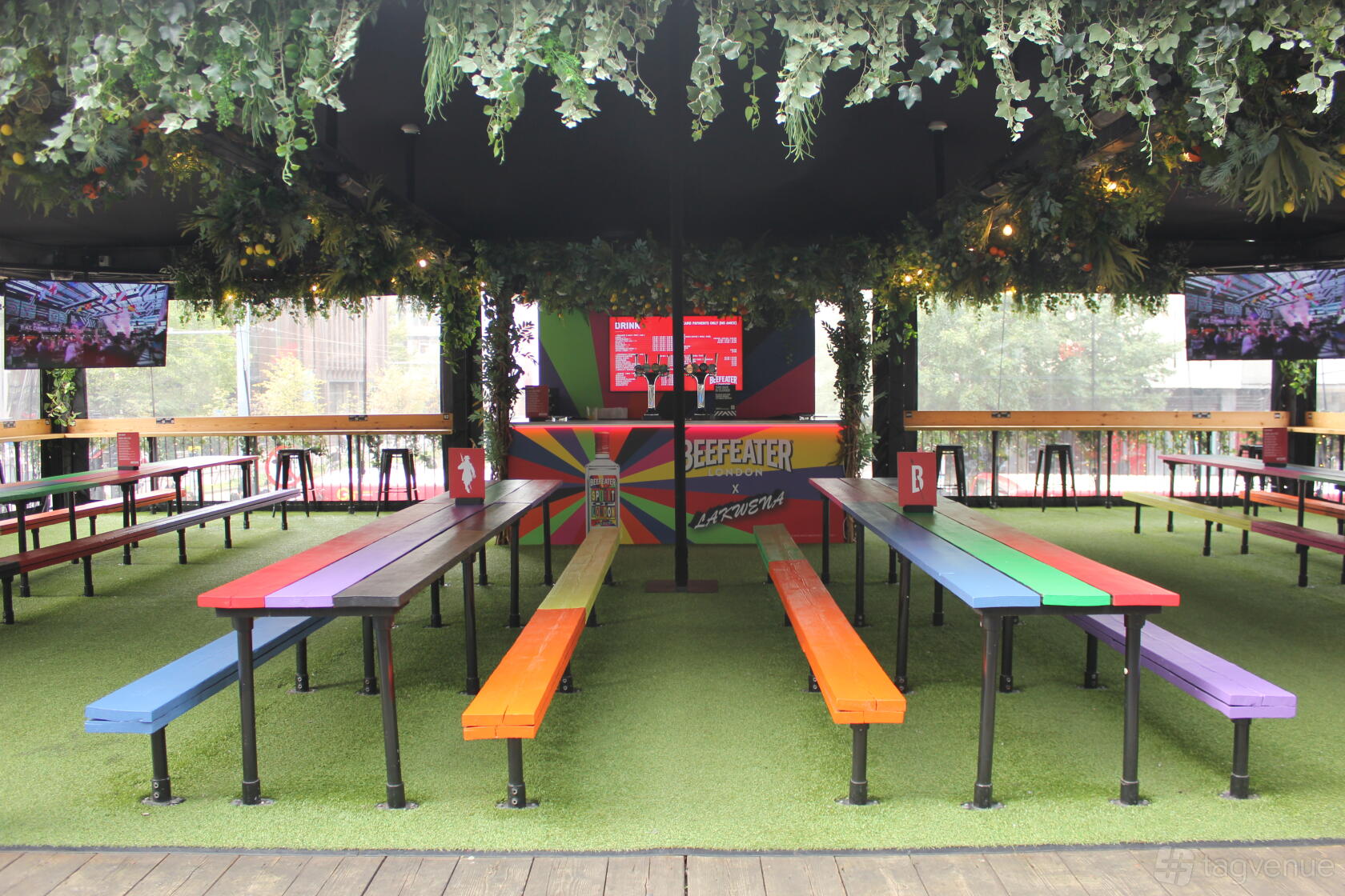 A restaurant terrace with rainbow-colored benches, artificial grass flooring, and overhead greenery at Boxpark Croydon.