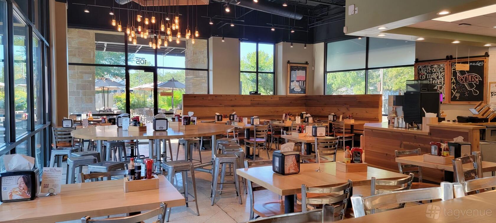 A restaurant dining area with wood tables, metal chairs, large windows, and a cluster of hanging lights at Maple Street Biscuit Company - Quarry Village.