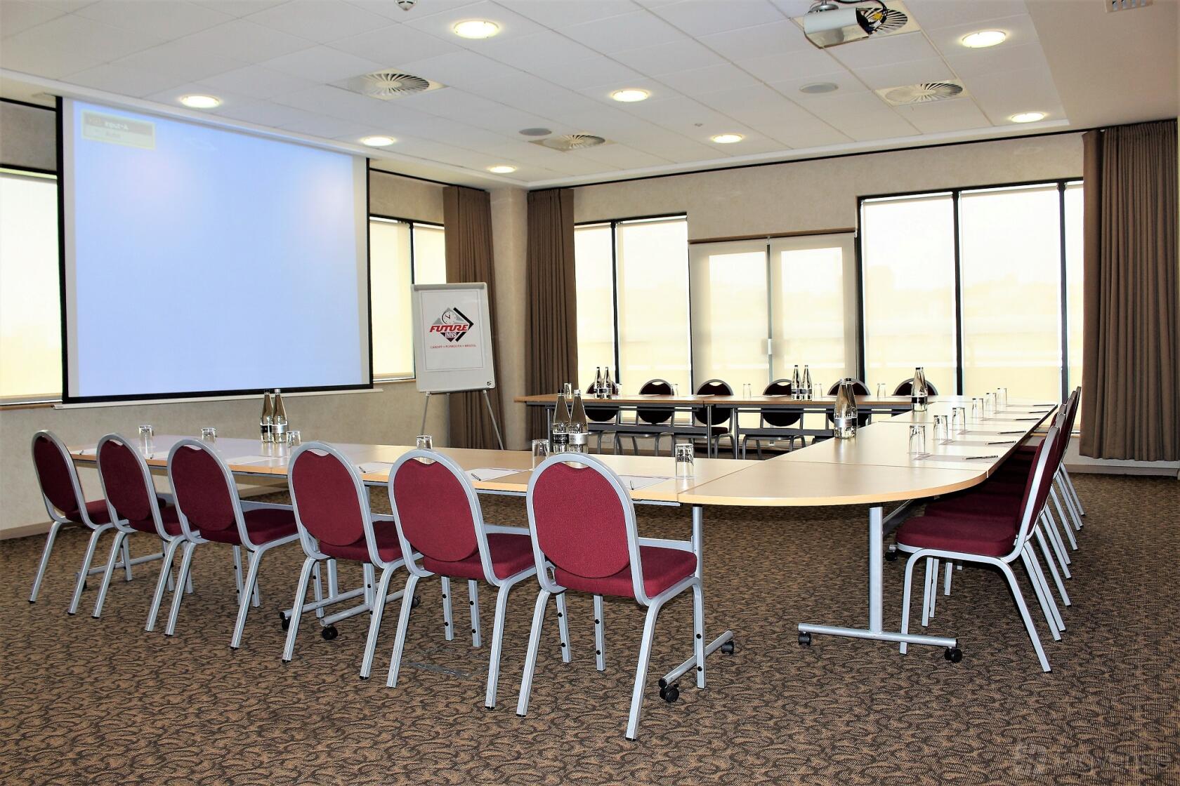A function room with U-shaped tables, red chairs, projection screen, and natural lighting at Future Inns Hotel Bristol.
