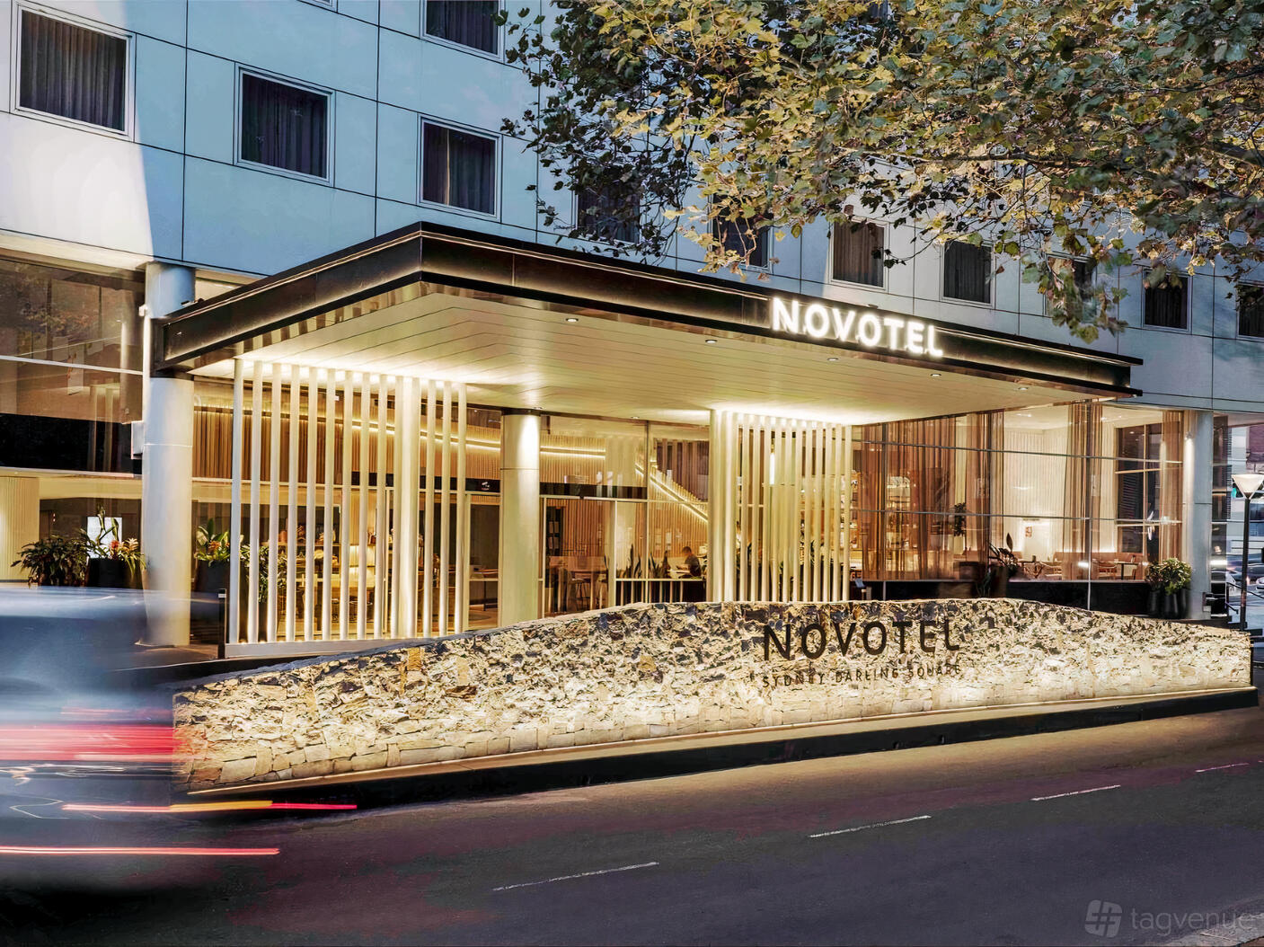 A hotel entrance with a stone feature wall, glass panels, and illuminated canopy at Novotel Sydney Darling Square.