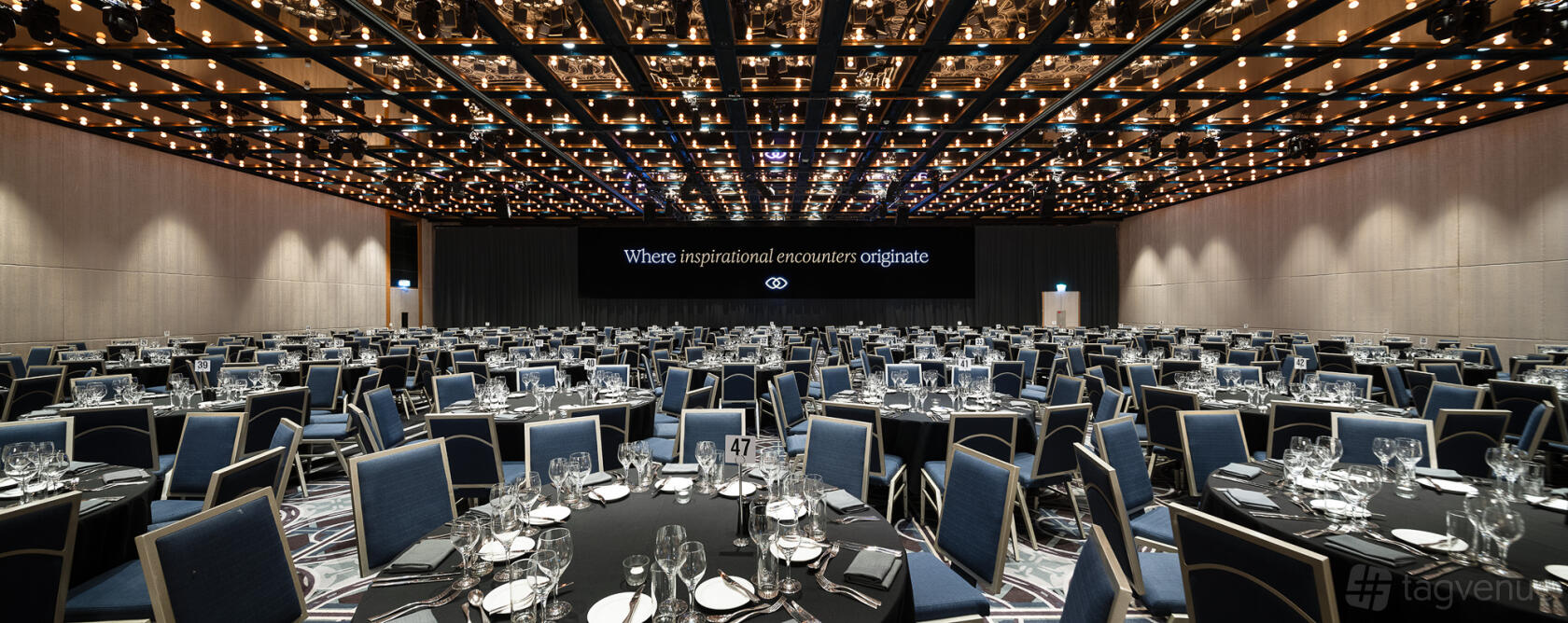 A hotel ballroom with a mirrored ceiling, banquet tables set with glassware, and a large presentation screen at Sofitel Sydney Wentworth.