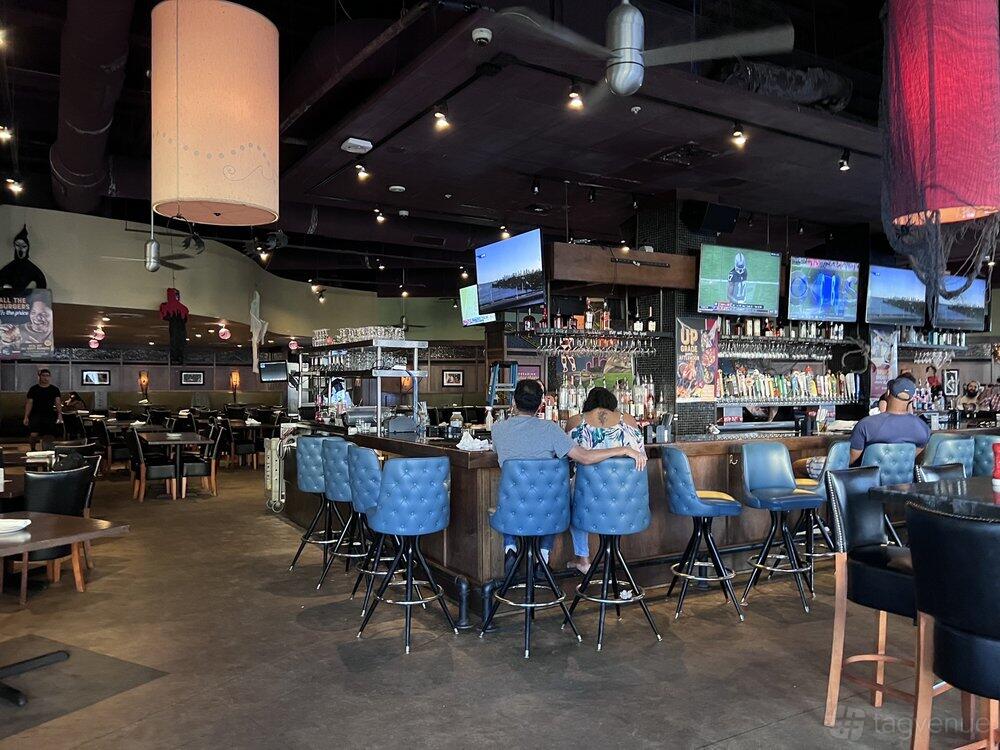 A bar with a long counter, blue barstools, multiple TVs, and a wall lined with beer taps at Bar Louie - Coconut Creek.