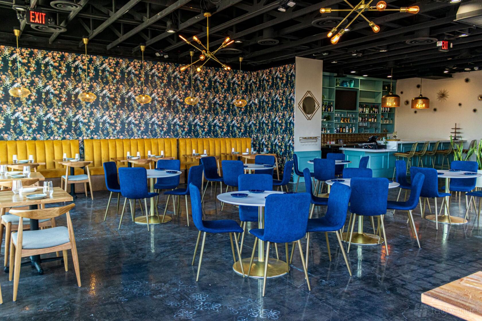A restaurant with blue velvet chairs, yellow tufted banquette seating, floral wallpaper, and gold light fixtures at Good Morning Kitchen and Cocktail Bar.