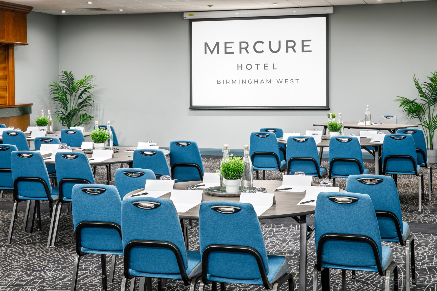 A hotel meeting room with blue chairs, tables set with notepads, and a projector screen at Mercure Birmingham West Hotel.