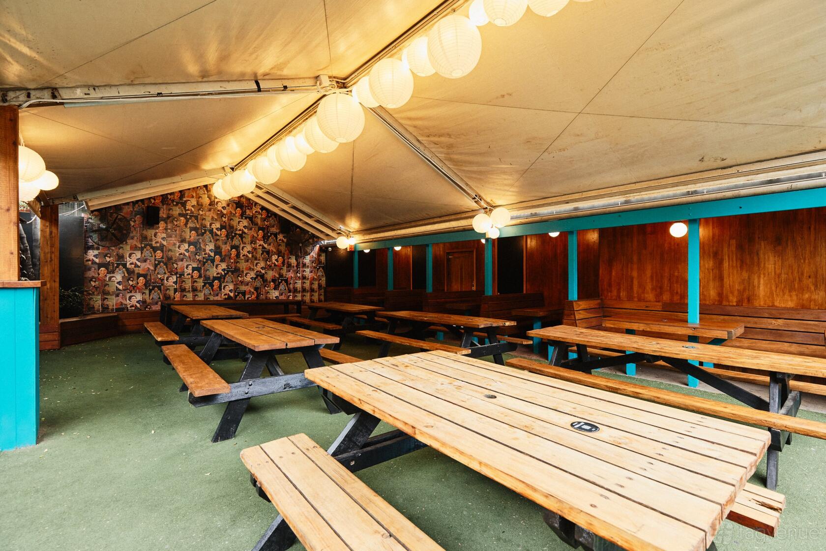 A pub space with wooden picnic tables, artificial grass flooring, and hanging lanterns at The Penny Black.