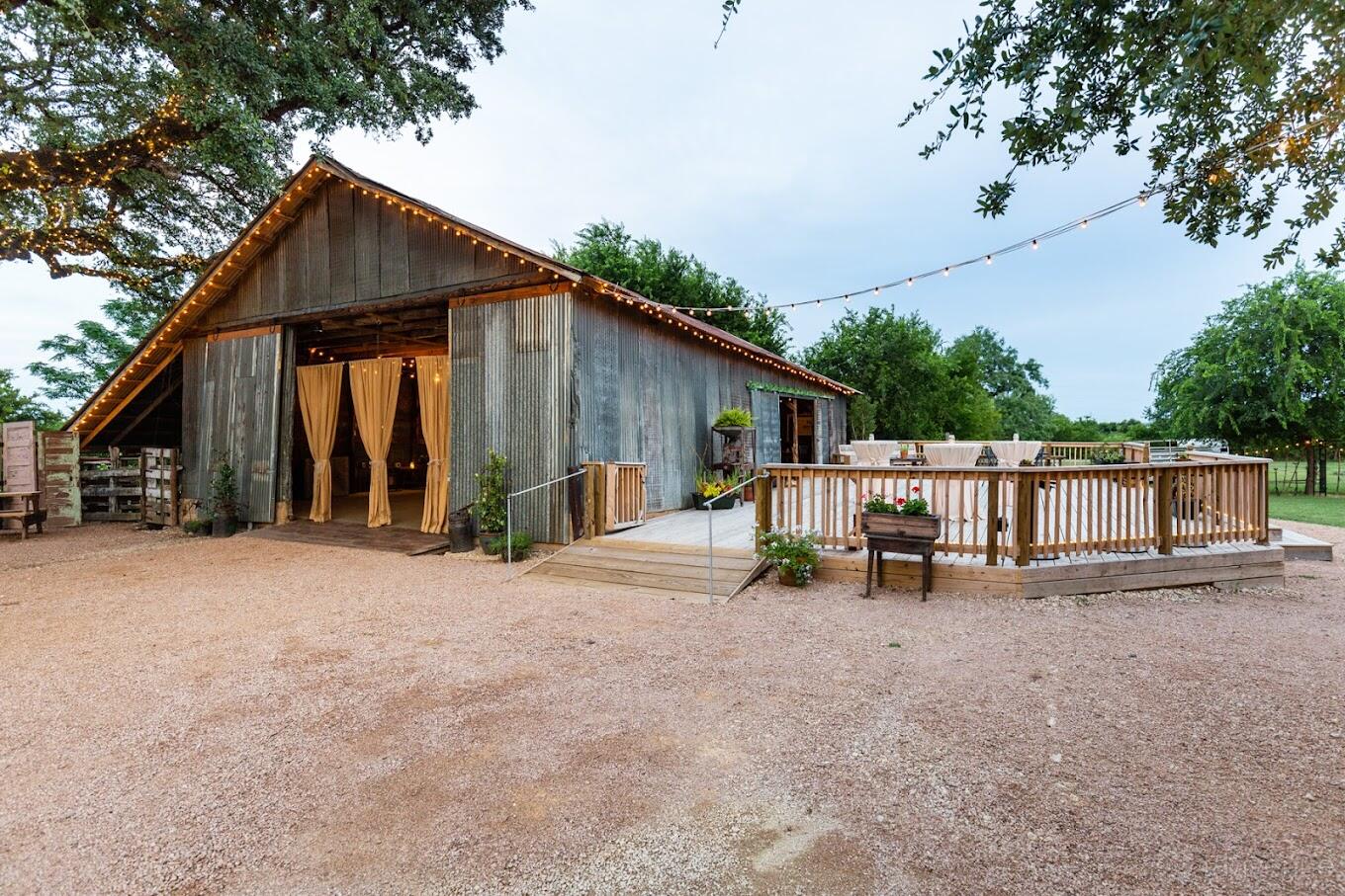 A rustic barn with weathered wood siding, string lights, and a wooden deck surrounded by open greenery at Texas Rock House.
