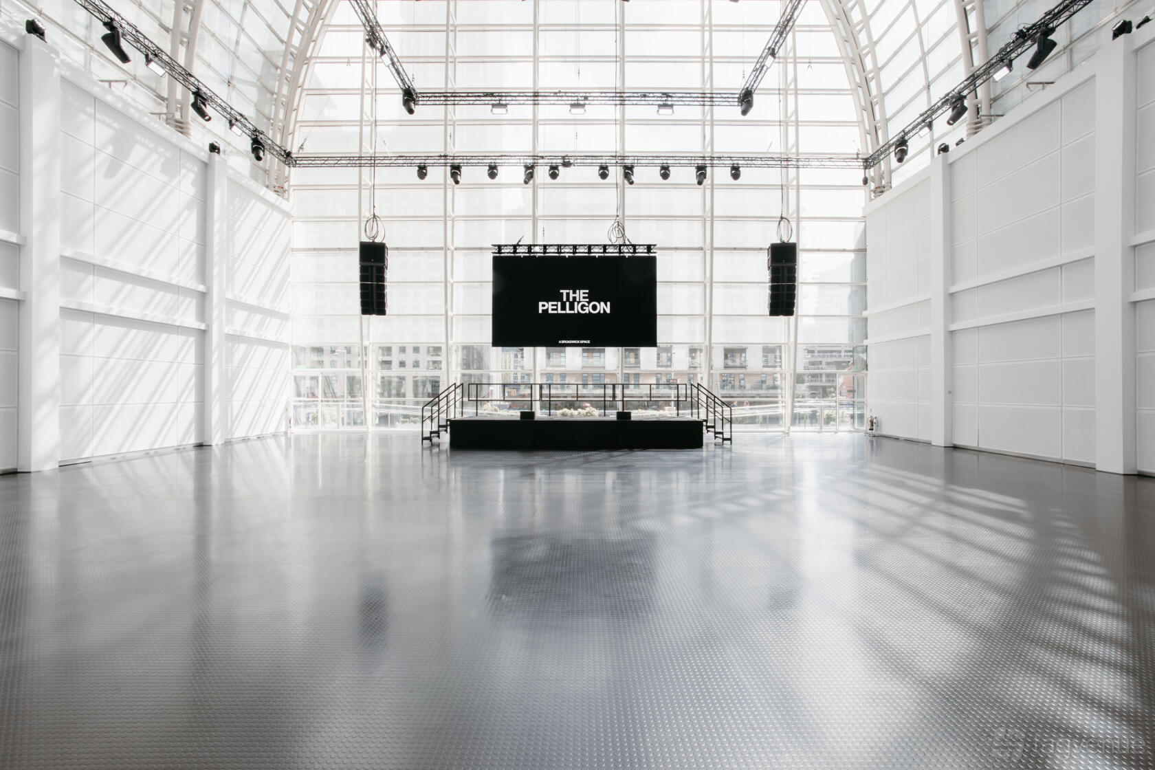 An event hall with floor-to-ceiling windows, exposed truss ceiling, and a stage with a large screen at The Pelligon.