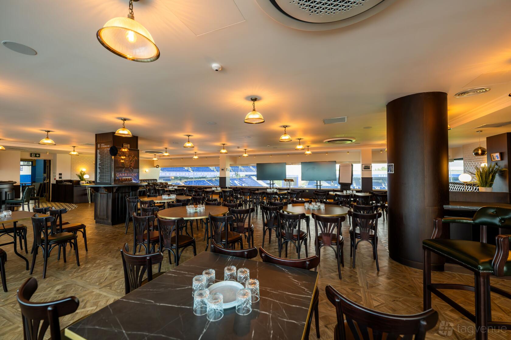 A function room with large windows overlooking the stadium, round tables, and wooden chairs at Birmingham City Football Club.