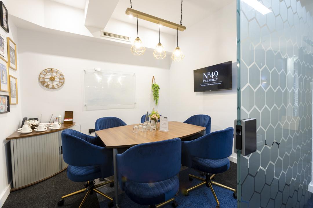 A meeting room with a wooden table, blue chairs, pendant lighting, and a wall-mounted screen at Biz Hub 49 Piccadilly.