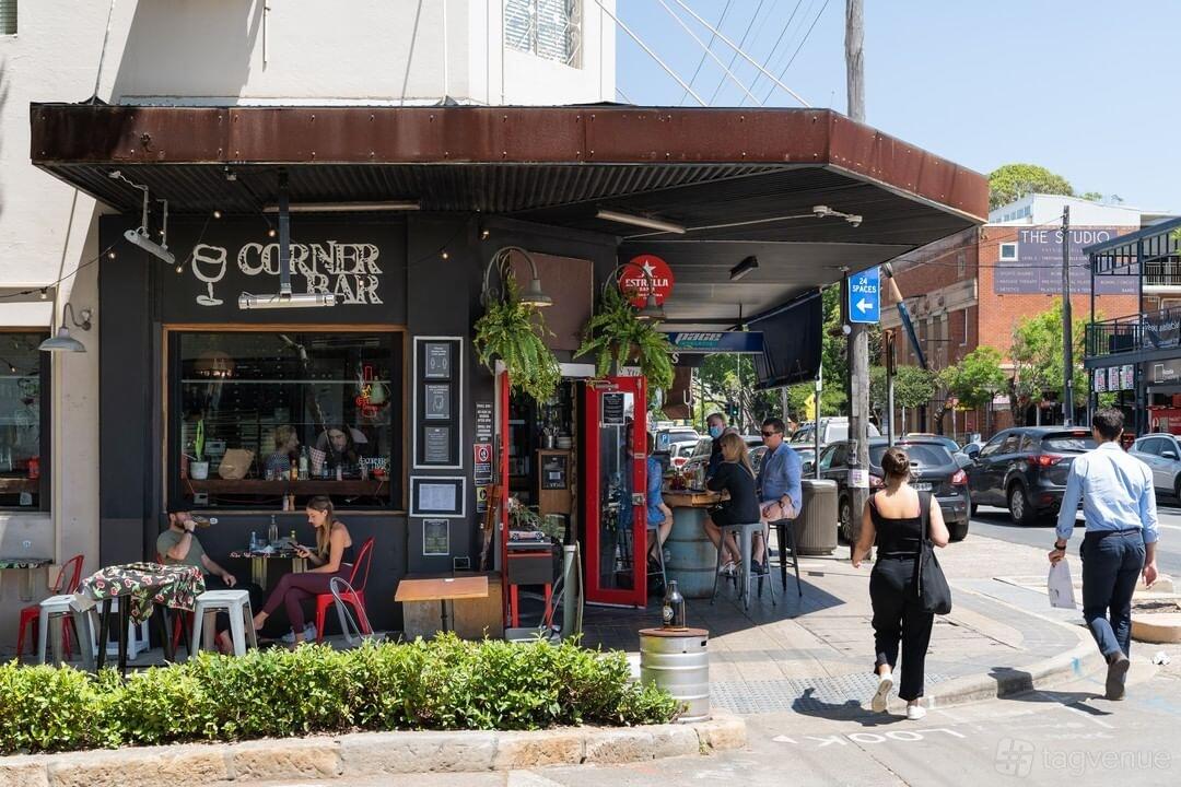 A street corner pub with outdoor tables, hanging plants, and a red entrance at Corner Bar Rozelle.
