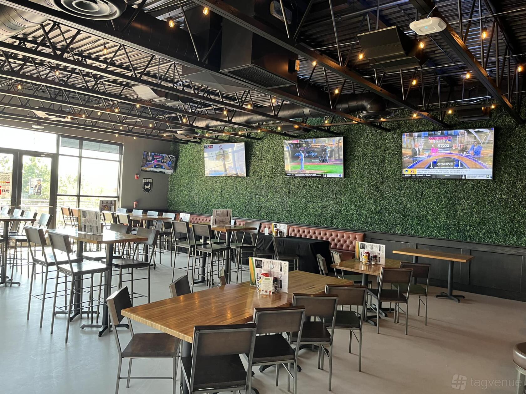 A bar with high-top wooden tables, wall-mounted TVs, and a green plant accent wall at Parlay Golf Sports Bar.