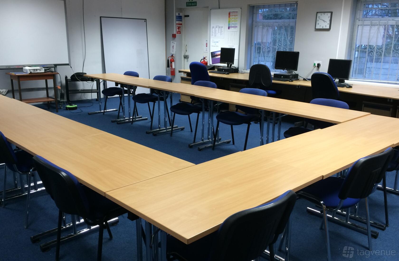 A university training room with U-shaped wooden tables, blue chairs, desktop computers, and large windows at Wesley House.