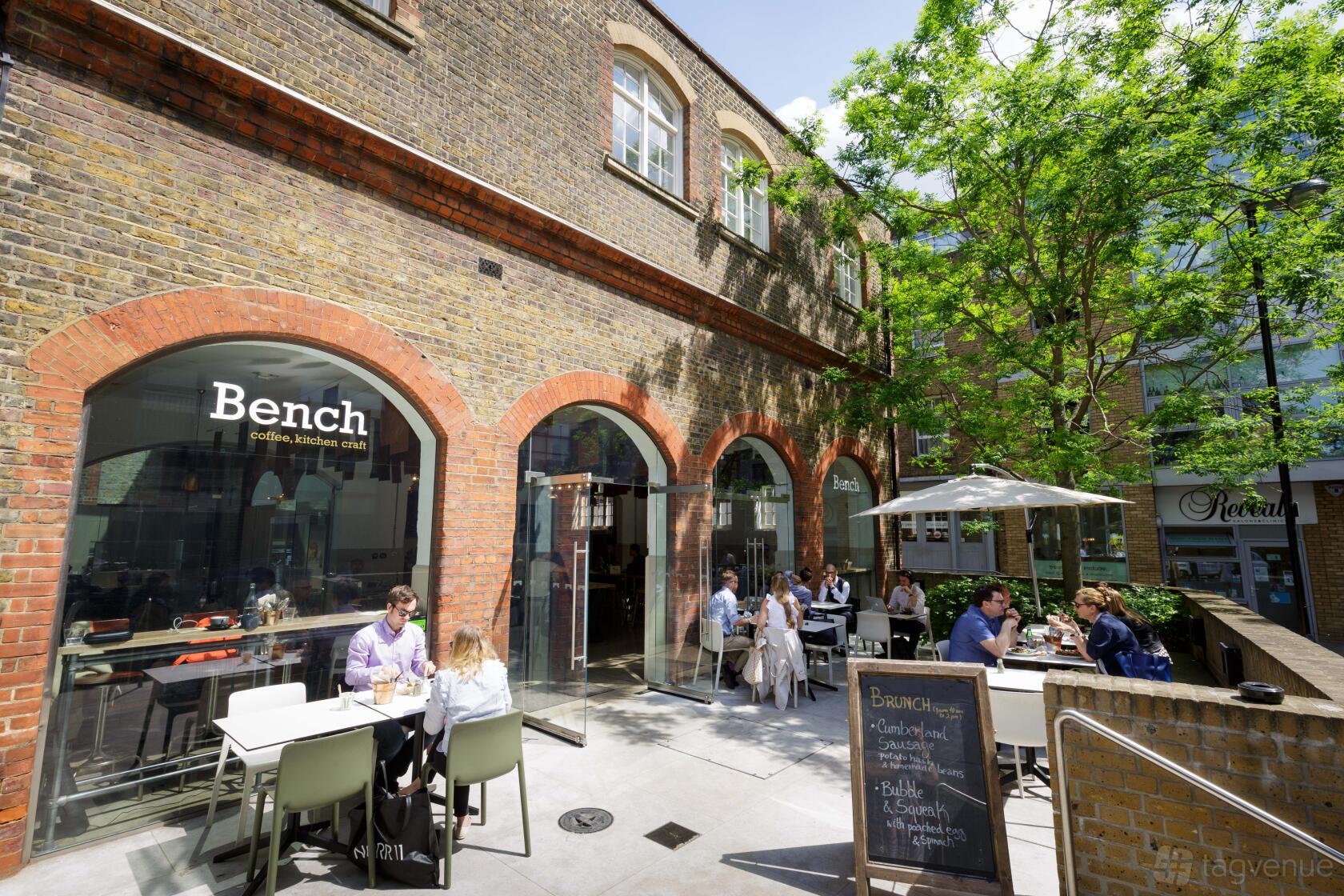 An outdoor event space with brick arches, glass doors, and patio seating beneath trees at The Goldsmiths' Centre.