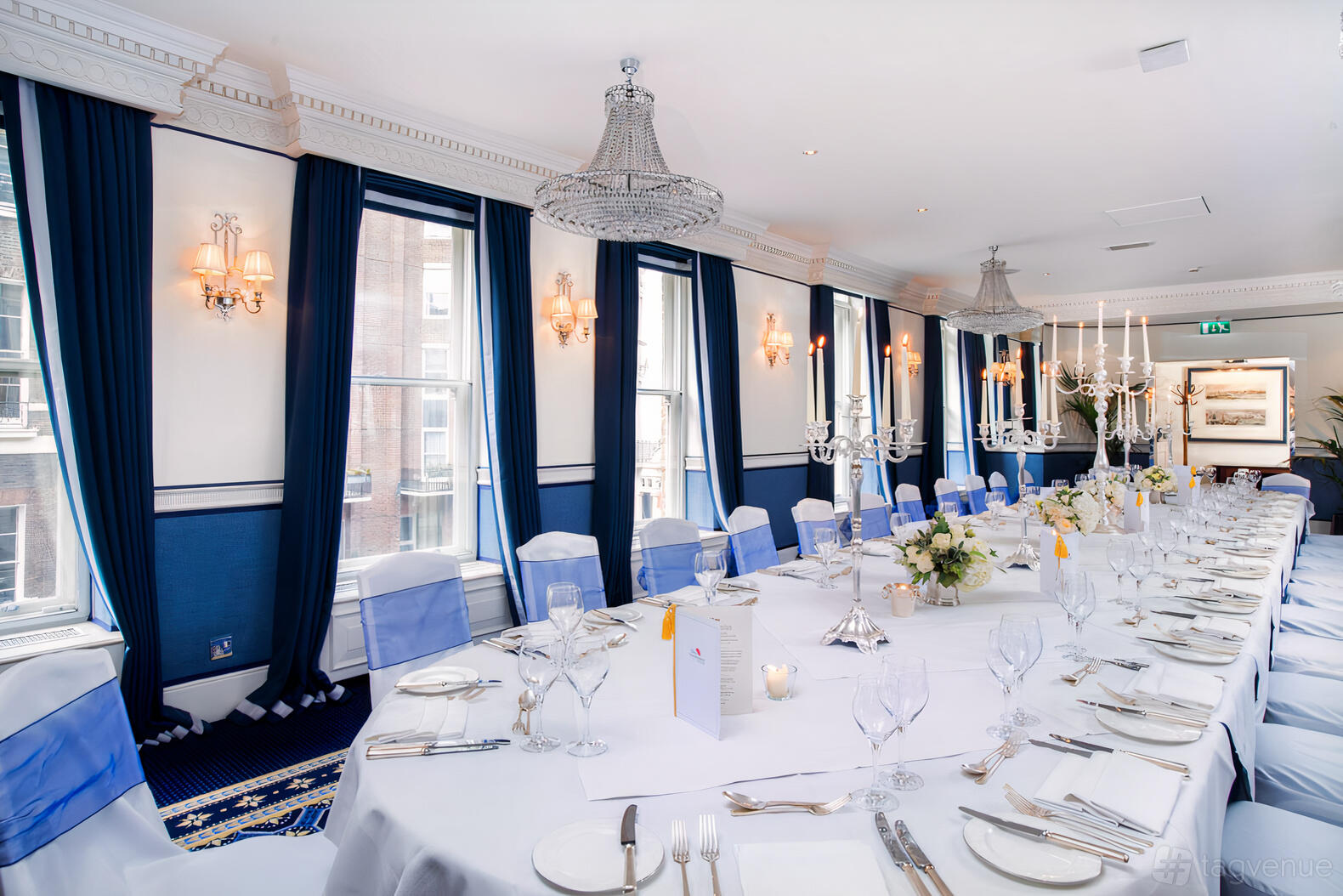 An event space with tall windows, blue curtains, chandeliers, and a long banquet table at The Chesterfield Mayfair.