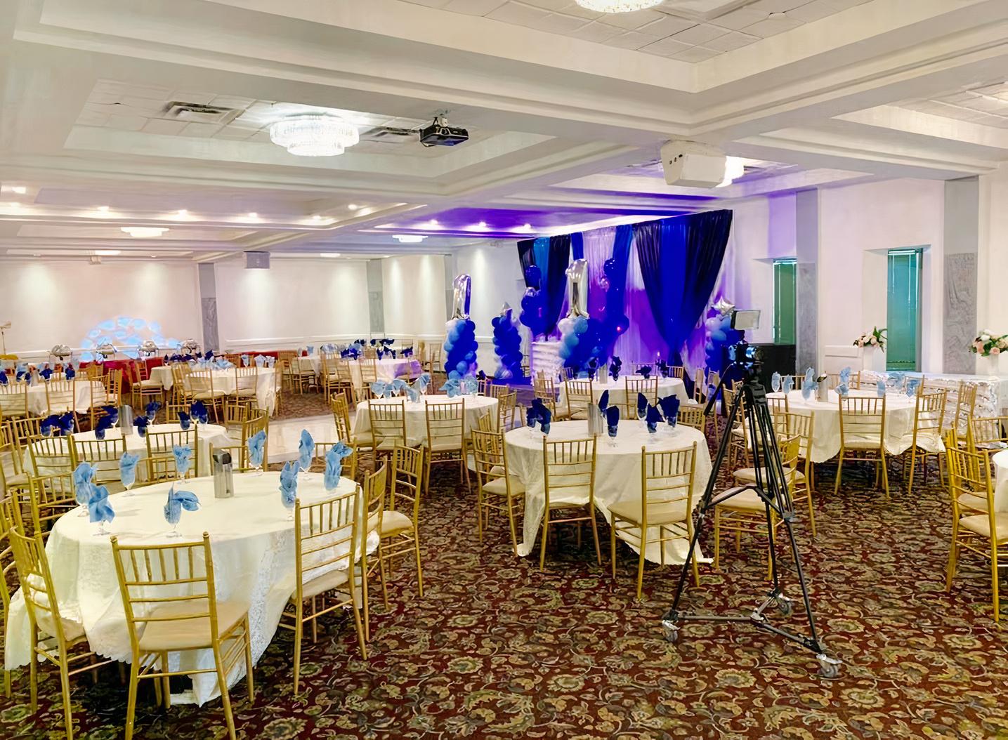 A banqueting hall with round tables dressed in white linens, gold chairs, and a decorated stage at Speranza Banquet Hall.