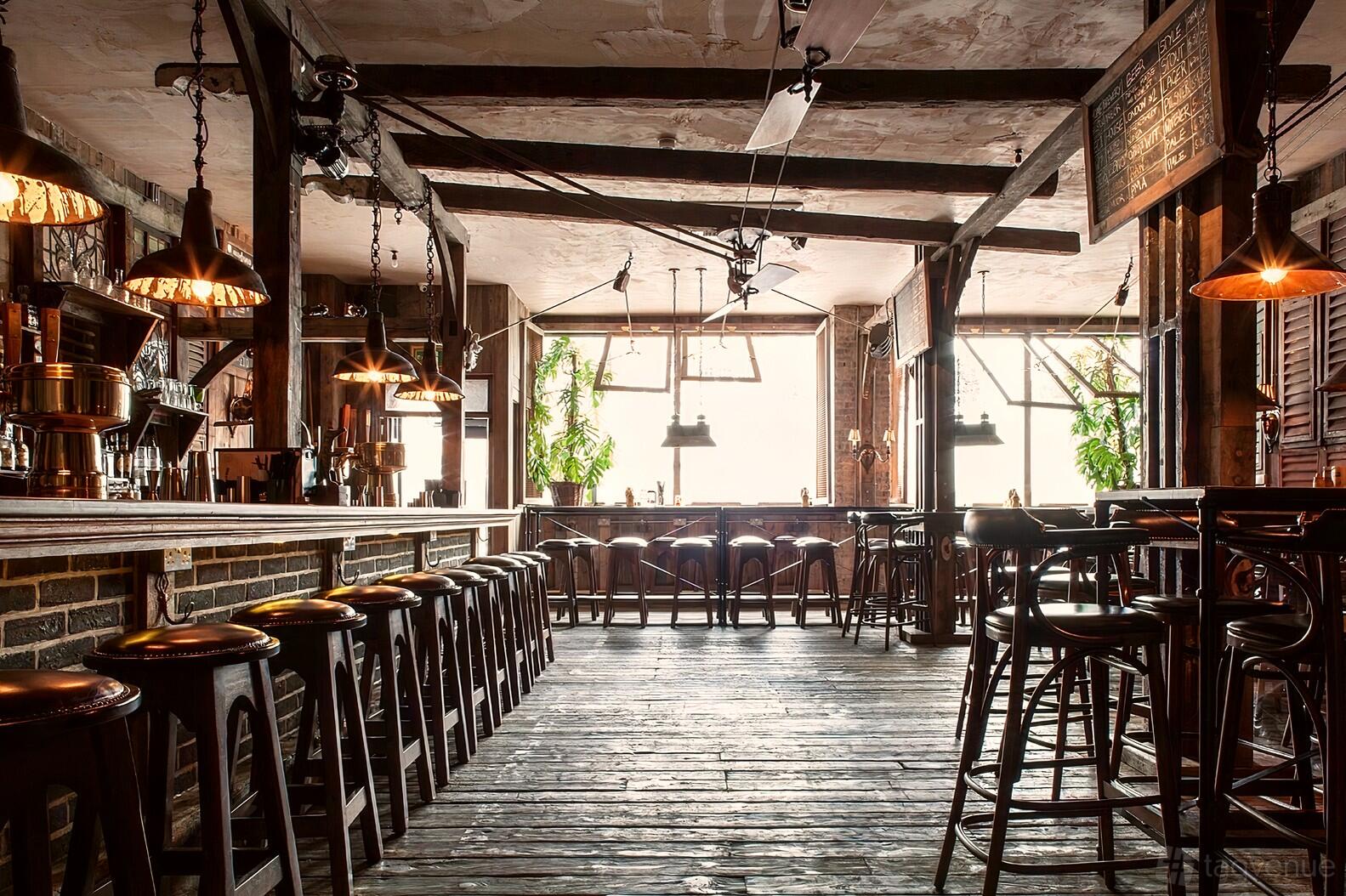 A bar with exposed wooden beams, rustic high stools, and large windows with natural light at Ninth Ward London.