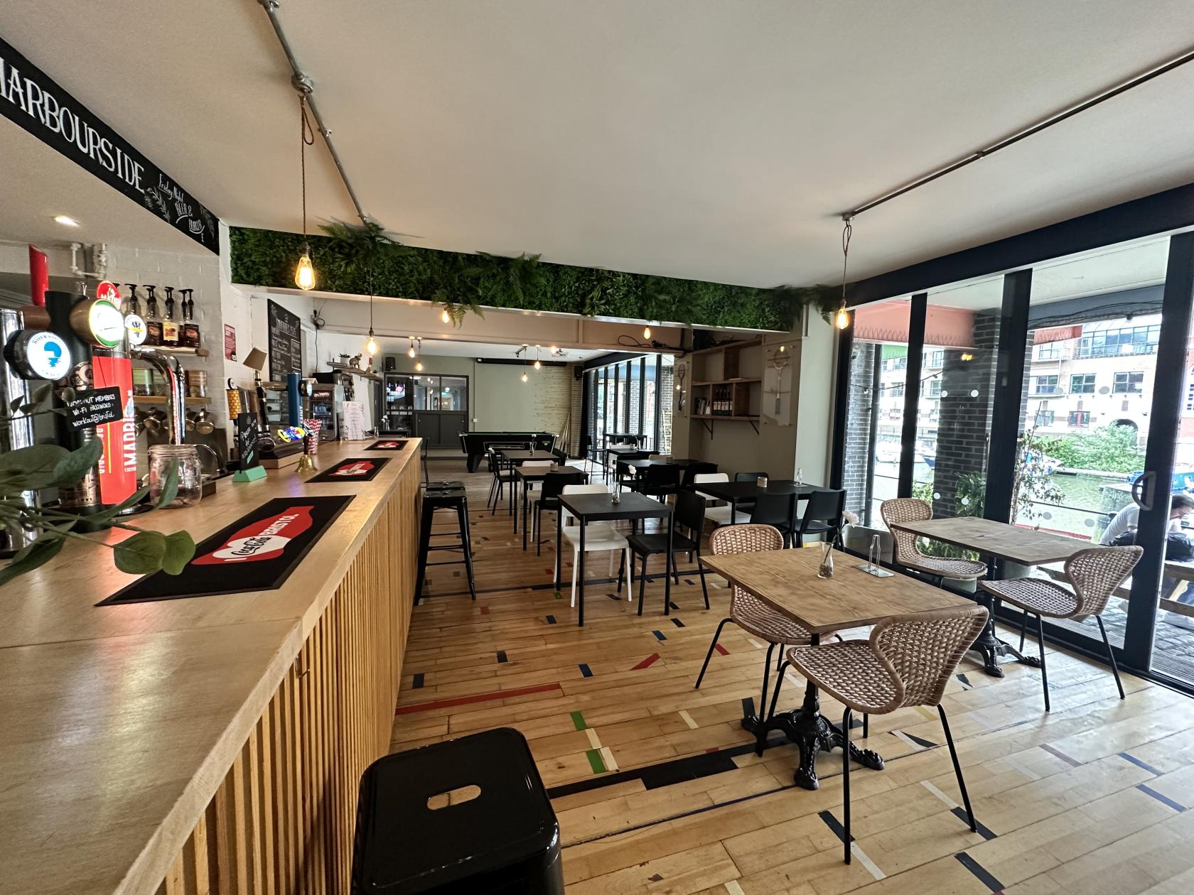 An event space in a bar with a long wooden counter, tap beers, black tables and chairs, and greenery above at Clubhaus Harbourside.
