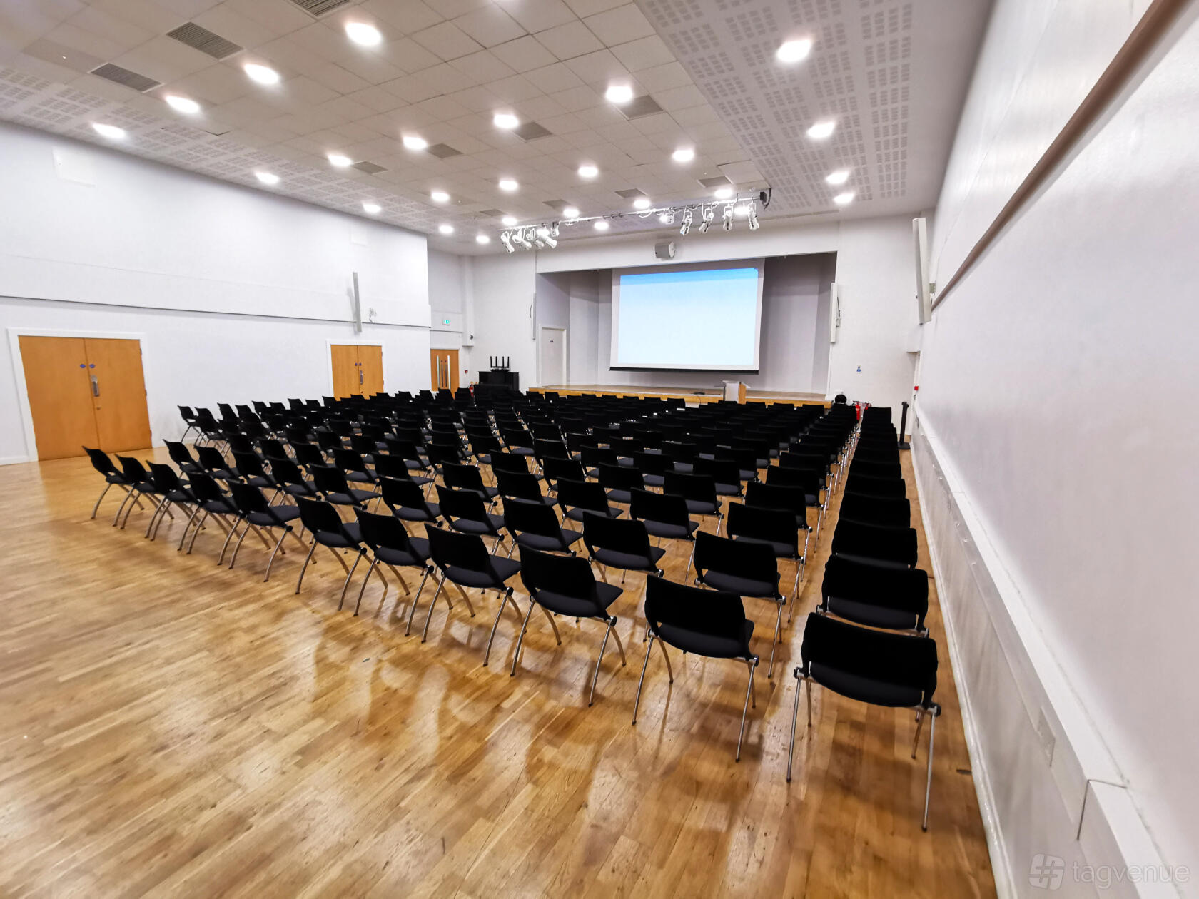 A hotel hall with rows of black chairs, parquet flooring, and a large projection screen at Queen's Gate House.