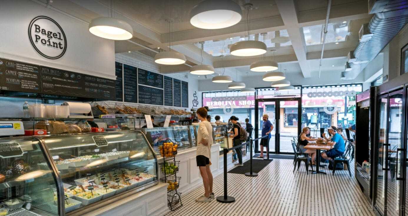 A restaurant with a glass pastry case, menu boards, and round ceiling lights at Bagel Point.