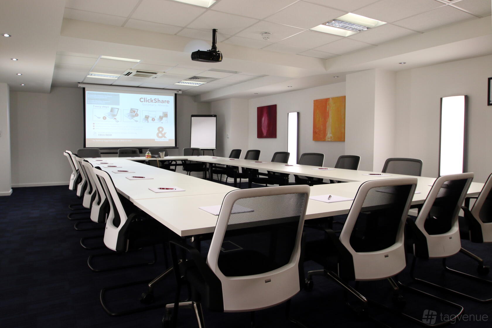 A meeting room with U-shaped white tables, ergonomic chairs, a ceiling projector, and wall art at 150 Minories.