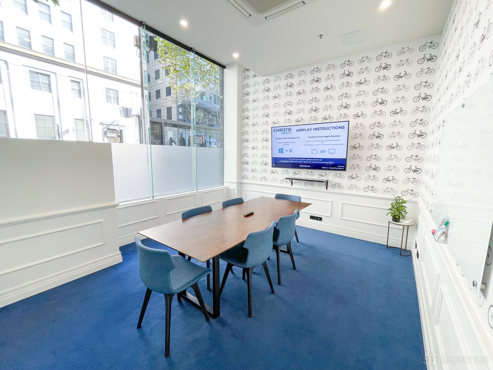A meeting room with a rectangular wooden table, blue chairs, wall-mounted screen, and bicycle-patterned wallpaper at Christie Spaces Conferencing Melbourne.
