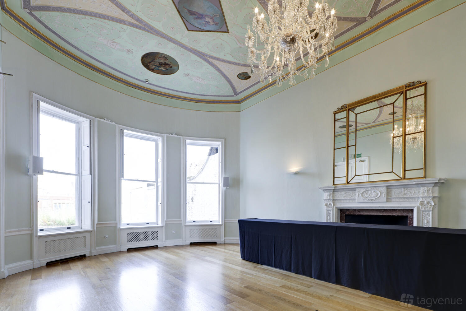 An event venue with tall bay windows, hardwood floors, a crystal chandelier, and ornate fireplace at Asia House.
