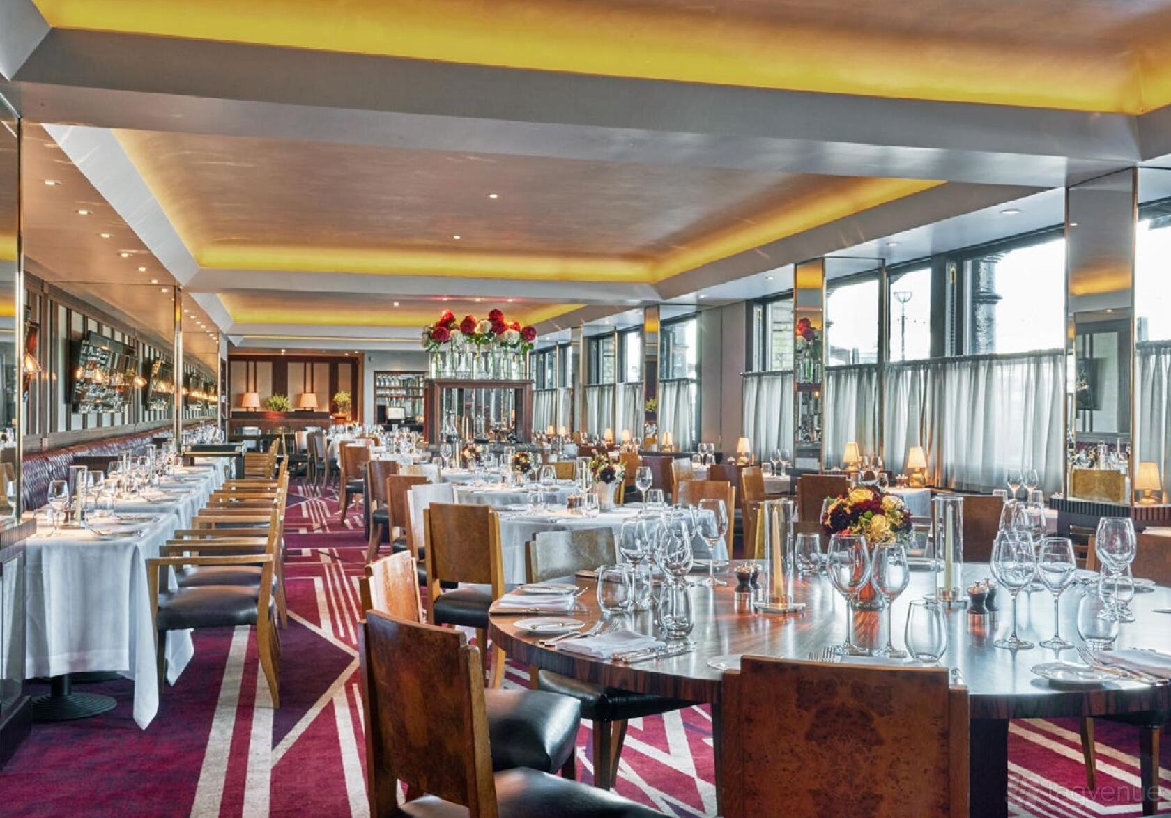 A restaurant with floor-to-ceiling windows, red carpet, and round tables set with glassware at Le Pont de la Tour.