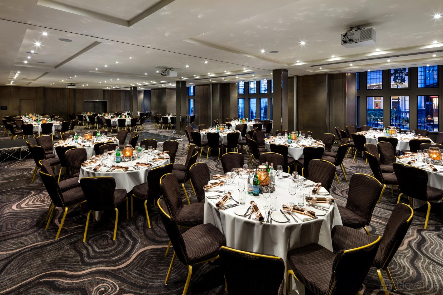 A hotel meeting room with round tables set with glassware, dark chairs, and patterned carpet at Radisson Blu Hotel, London Bloomsbury.