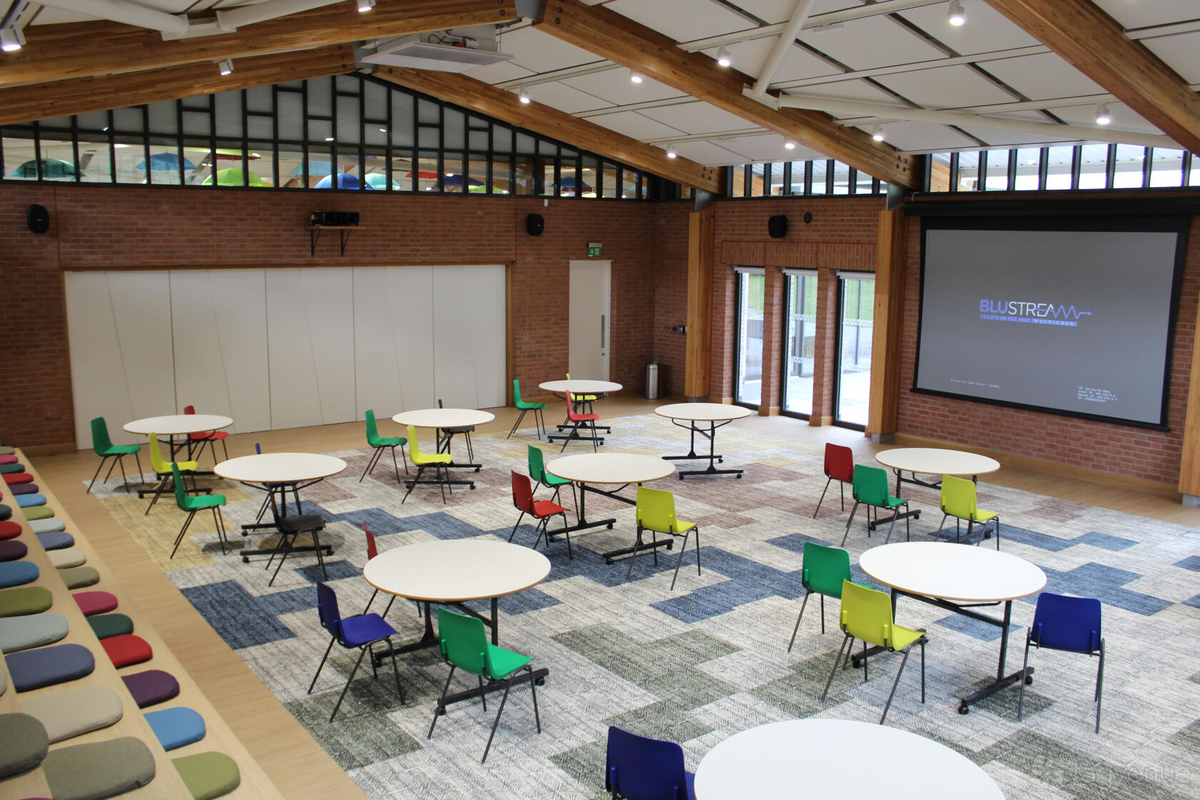 An event space with round tables, colorful chairs, tiered seating, brick walls, and a projector screen at The Nest.