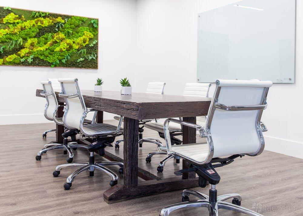 A boardroom with a rustic wooden table, white leather chairs, a glass whiteboard, and a green wall feature at Maketory.