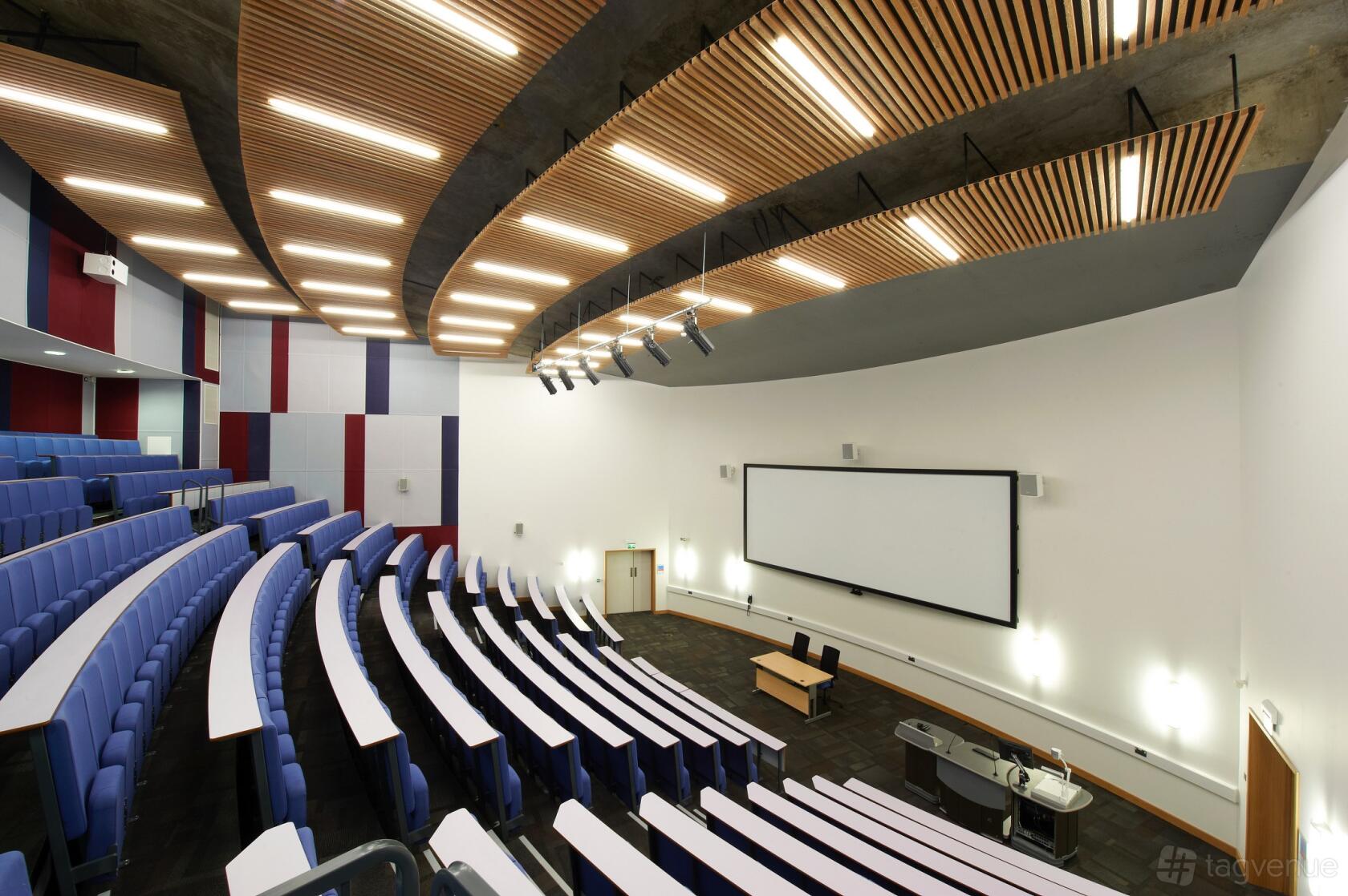 An auditorium with tiered seating, curved wood ceiling panels, and a large projection screen at ARU Venue Hire - Cambridge.