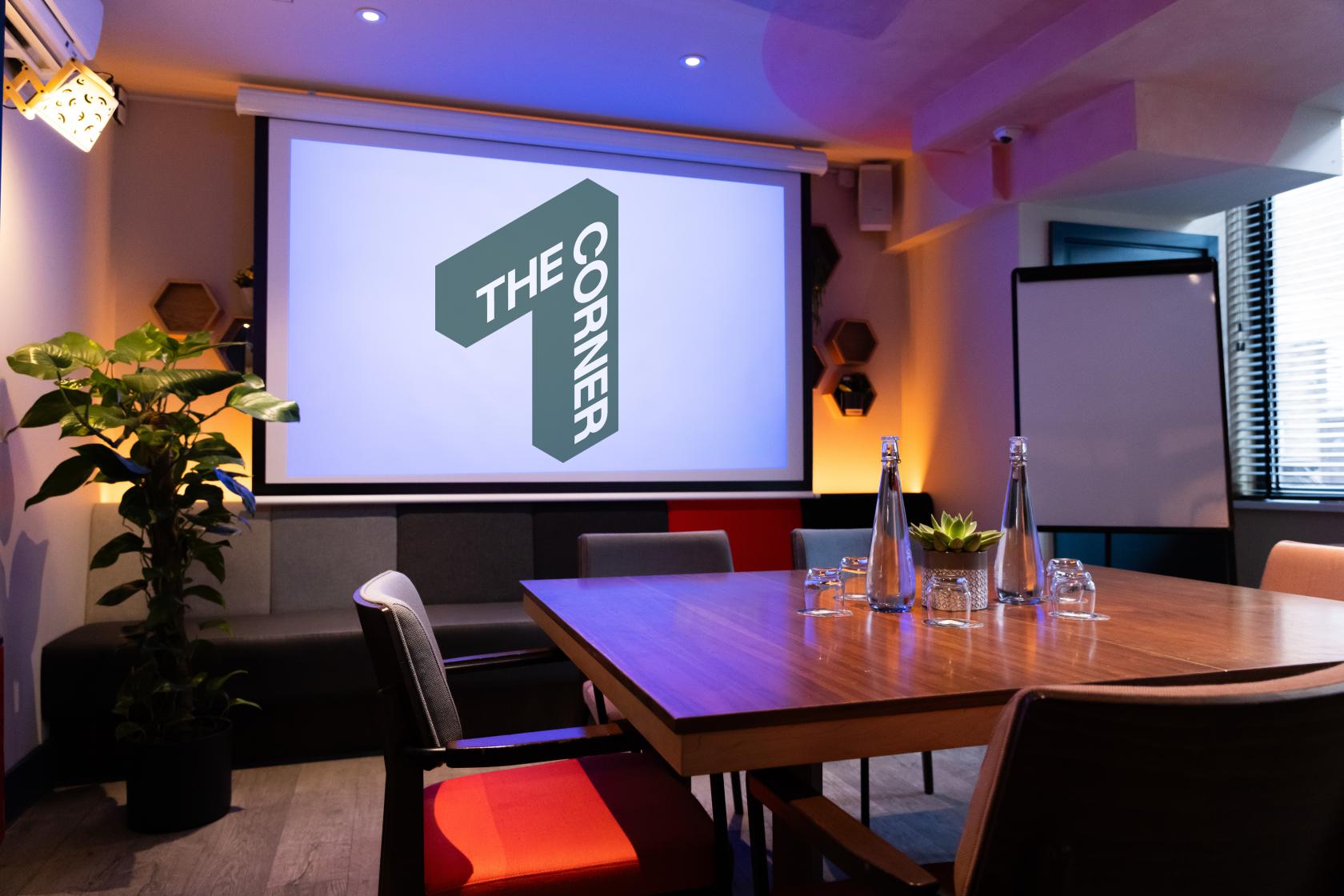 A meeting room with a wooden conference table, projector screen, flip chart, and potted plants at The Corner London City.