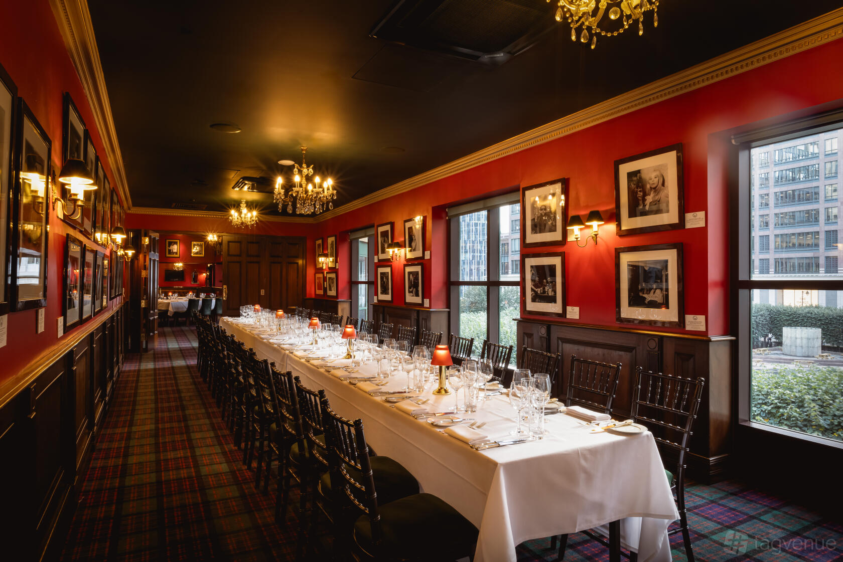 A dining room with a long table set with white linens, dark wood chairs, and red walls adorned with framed photos at Boisdale of Canary Wharf.